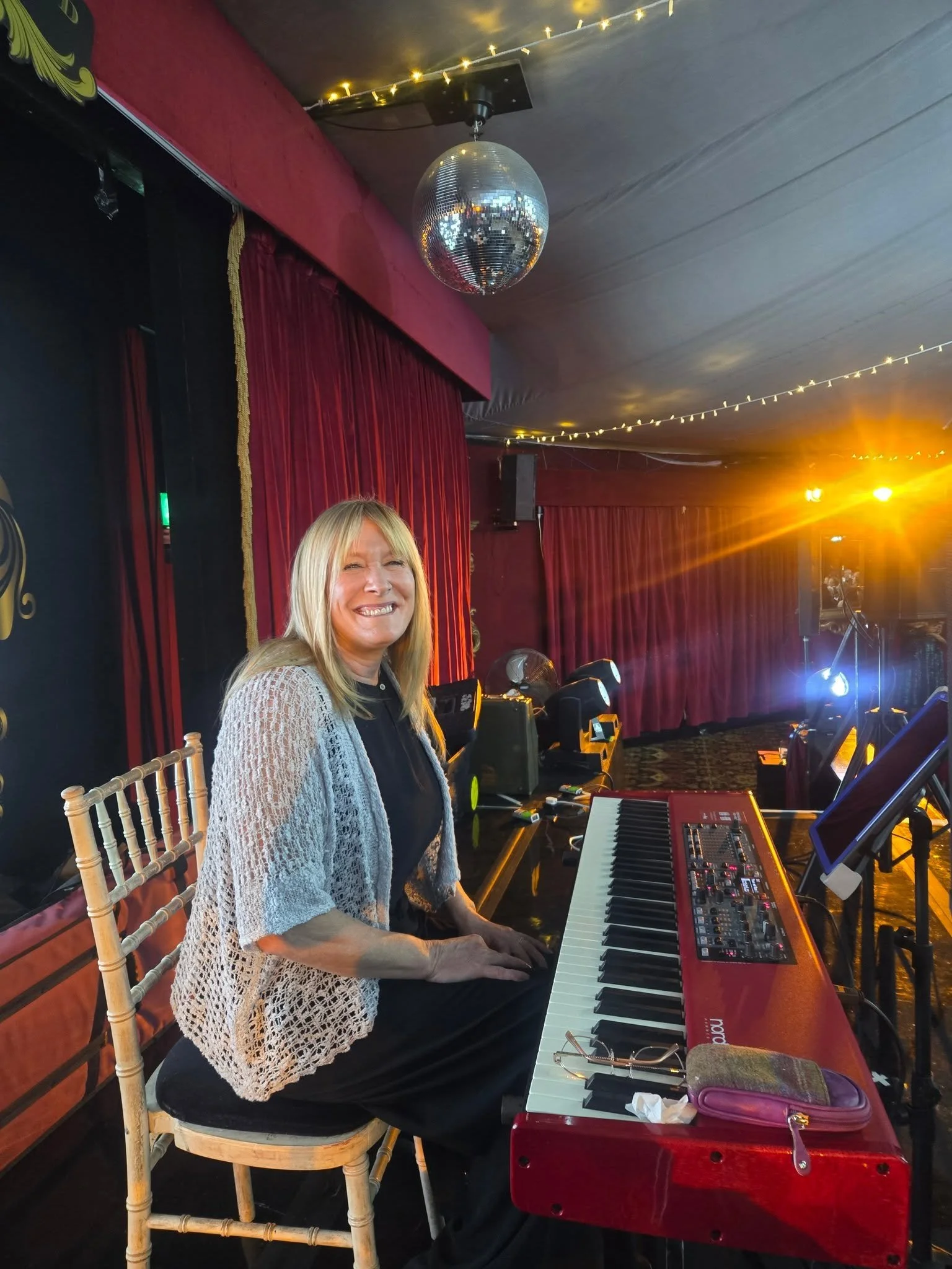 A woman playing a red keyboard on stage near a red curtain, with a disco ball hanging from the ceiling and stage lights in the background. Clappy Doo Ceilidh band Scottish Ceilidh band Scottish music