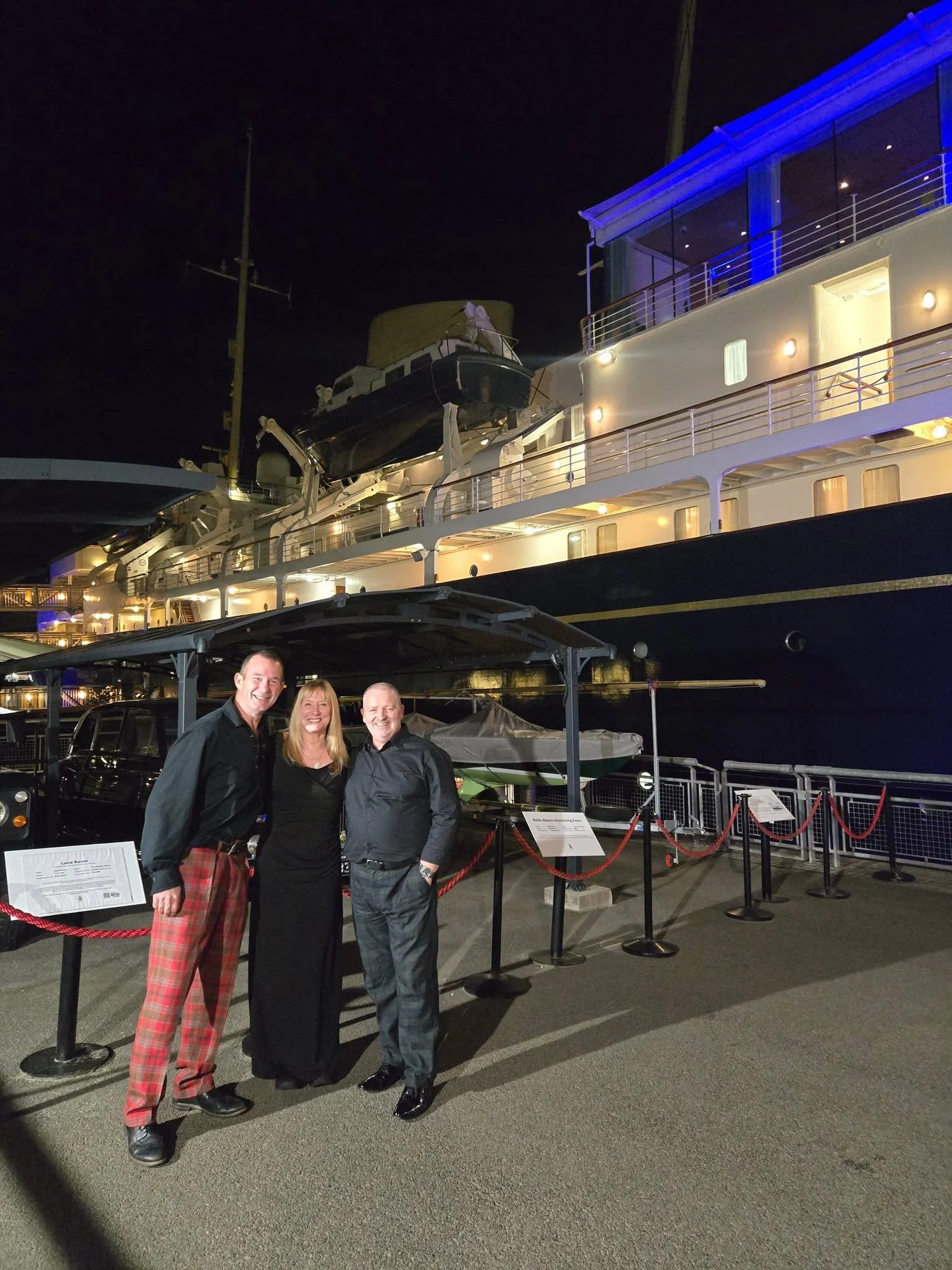 Three people stand in front of a large yacht at night. The yacht is illuminated with several lights and has balconies and a lifeboat on the side. The three people are smiling and dressed in dark, formal attire.