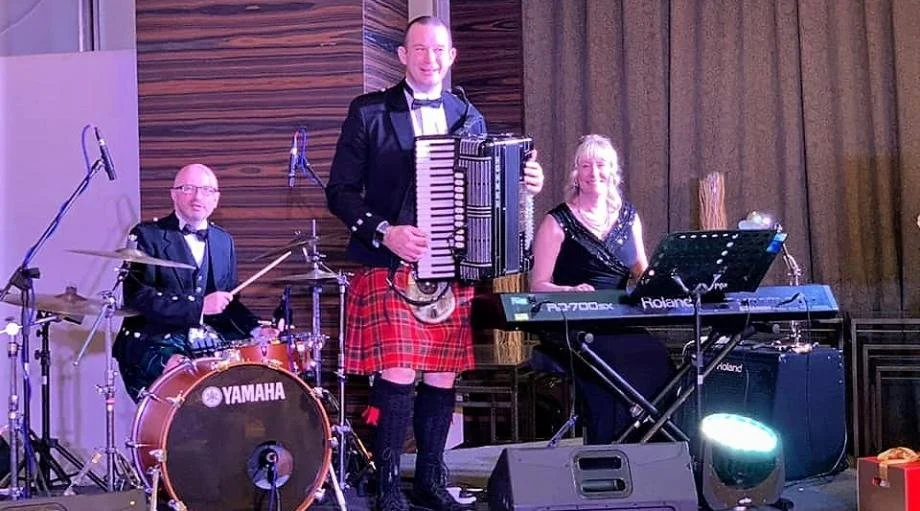 Three musicians performing on stage: a drummer on the left, a man in the center playing an accordion and wearing a kilt, and a woman on the right playing a keyboard, all dressed in formal attire.