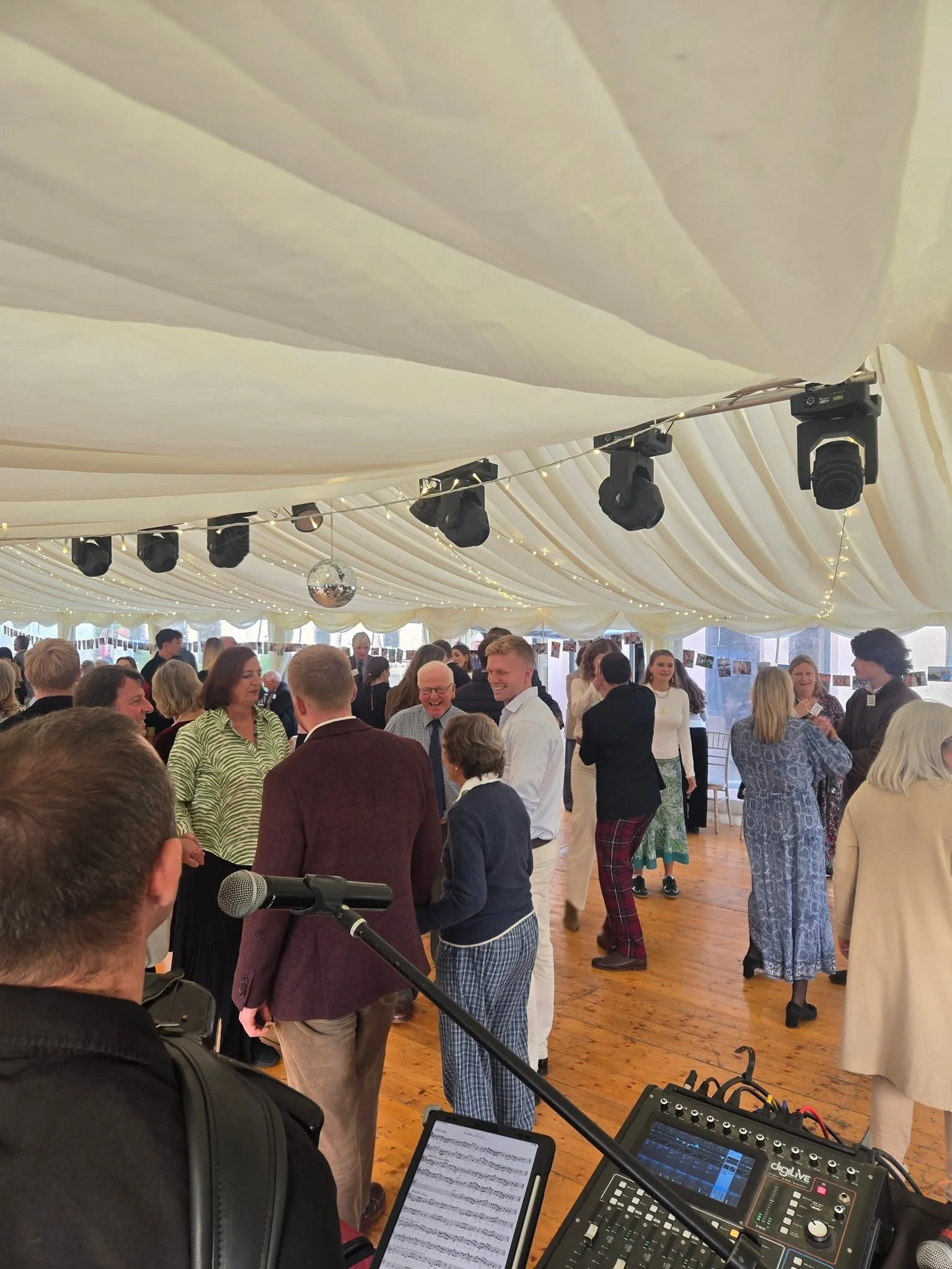 People attending a social event or celebration in a large tent with string lights and a disco ball, engaging in conversations and smiling.  Clappy Doo Ceilidh band Scottish Ceilidh band Scottish music