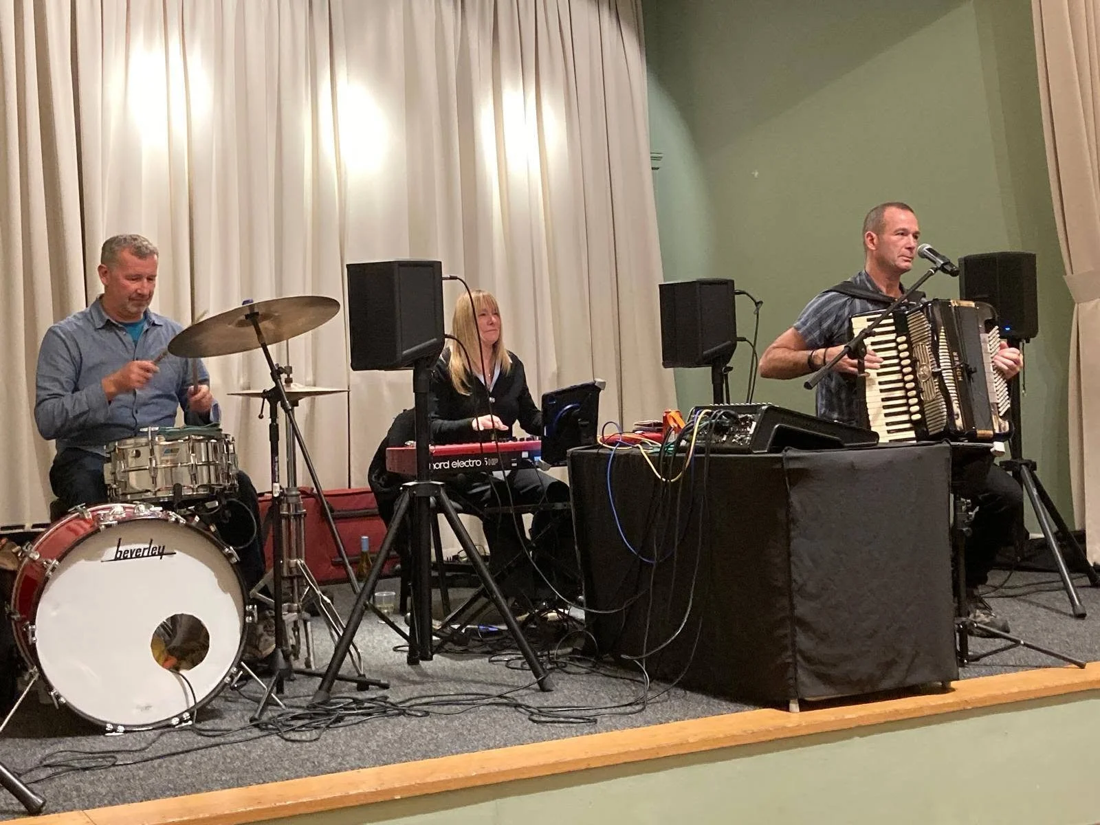 Three musicians performing on stage: a man playing drums, a woman on keyboard, and a man playing accordion.  Clappy Doo Ceilidh band Scottish Ceilidh band Scottish music