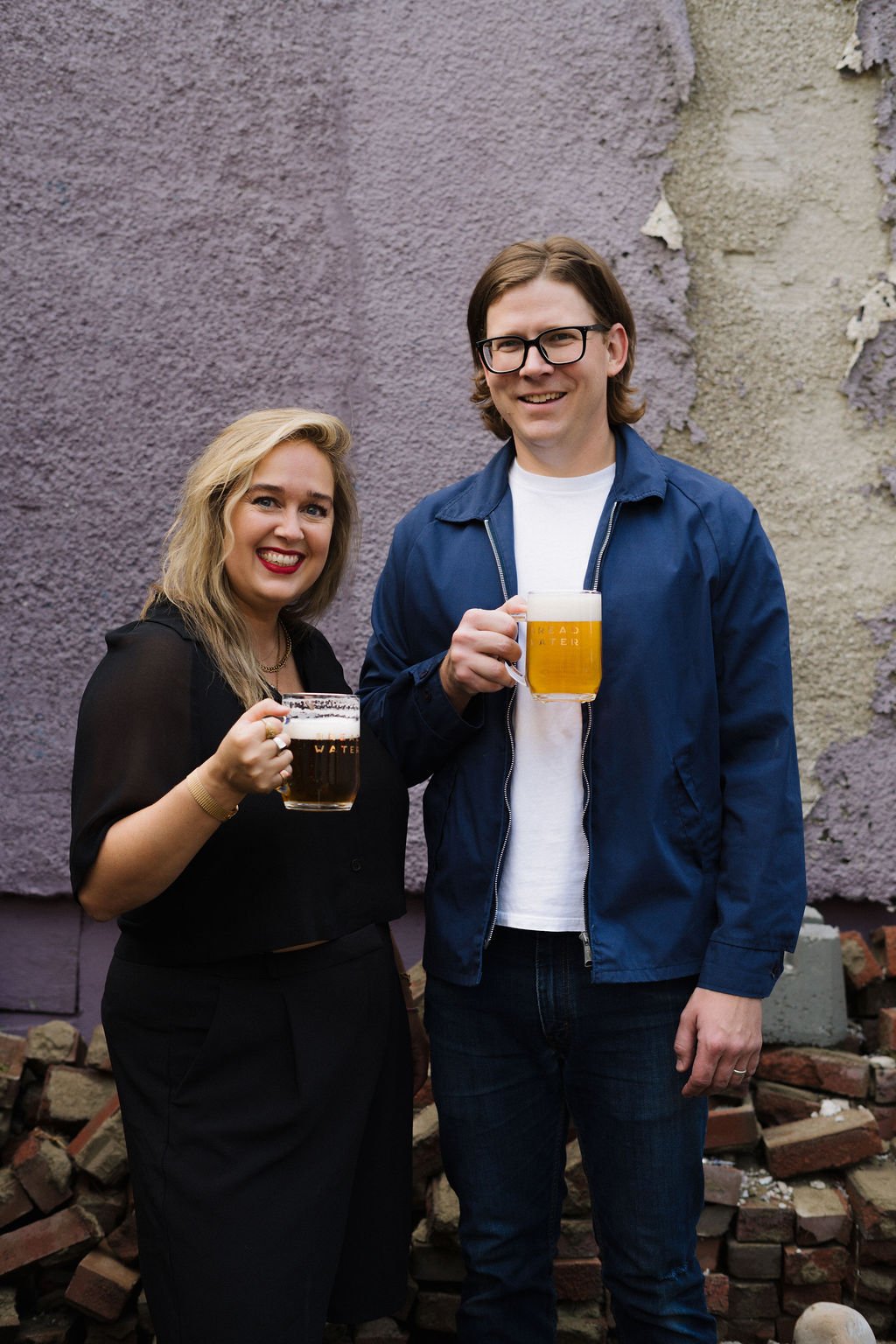 Blake and Alexa of Breadwater Beer smiling and holding mugs of beer, standing outside against a textured wall with peeling paint and scattered bricks.
