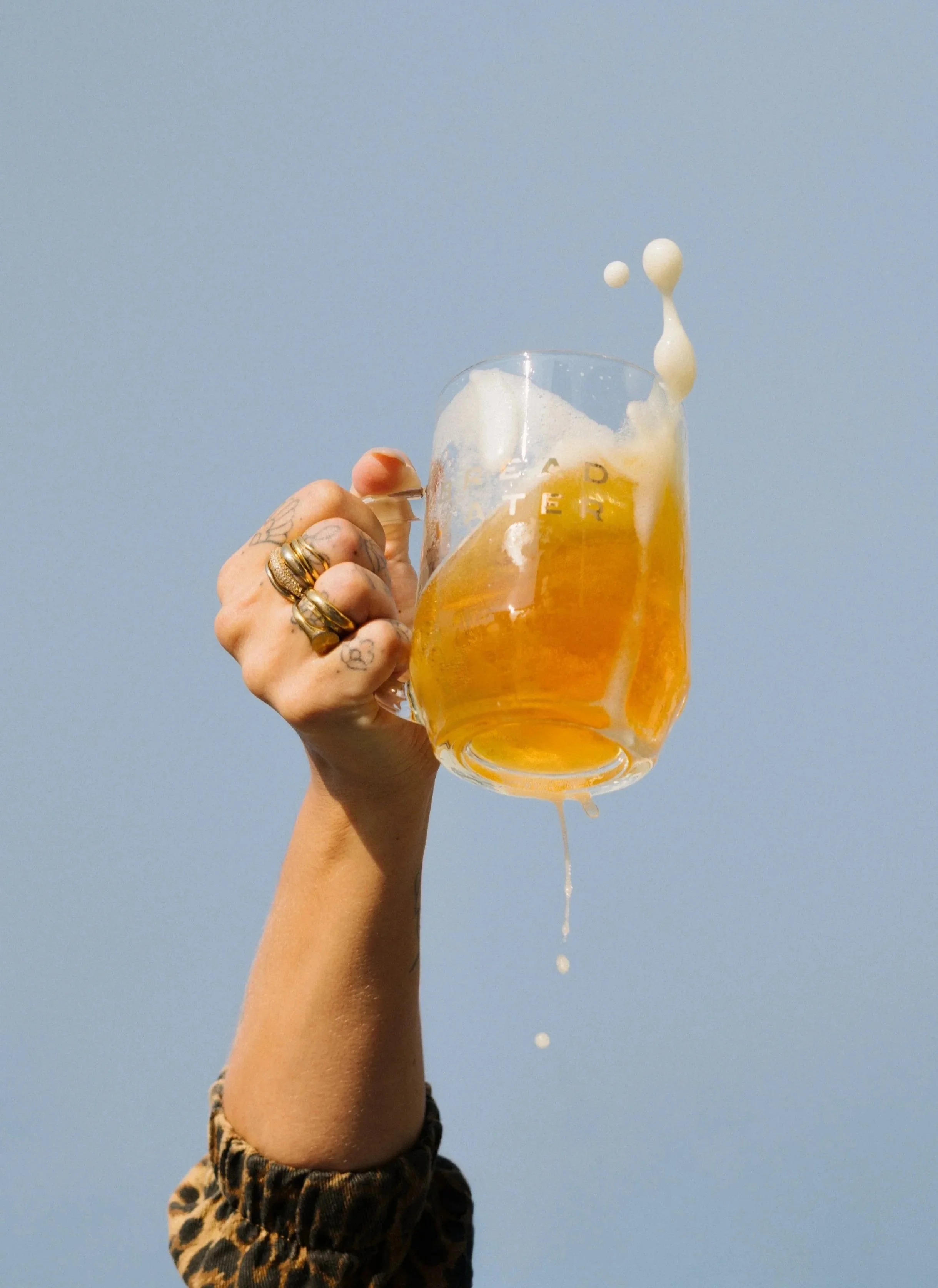 A person's hand holding a glass mug of Bread Water beer against a clear blue sky, with a splash of foam spilling from the mug, and droplets falling down.