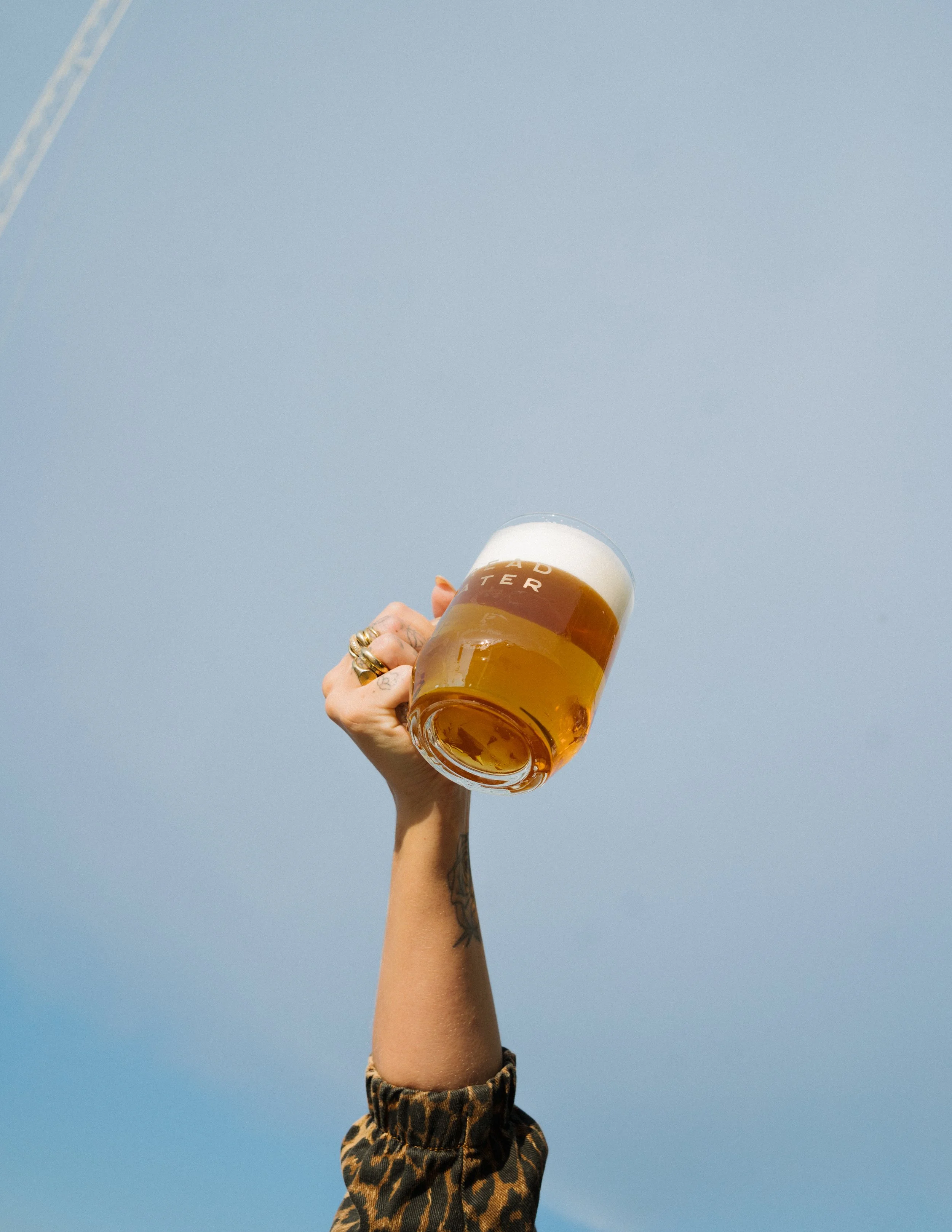 A woman raises her arm holding a glass of Bread Water beer in the air against a clear blue sky. She is wearing gold rings and a patterned long-sleeve top.