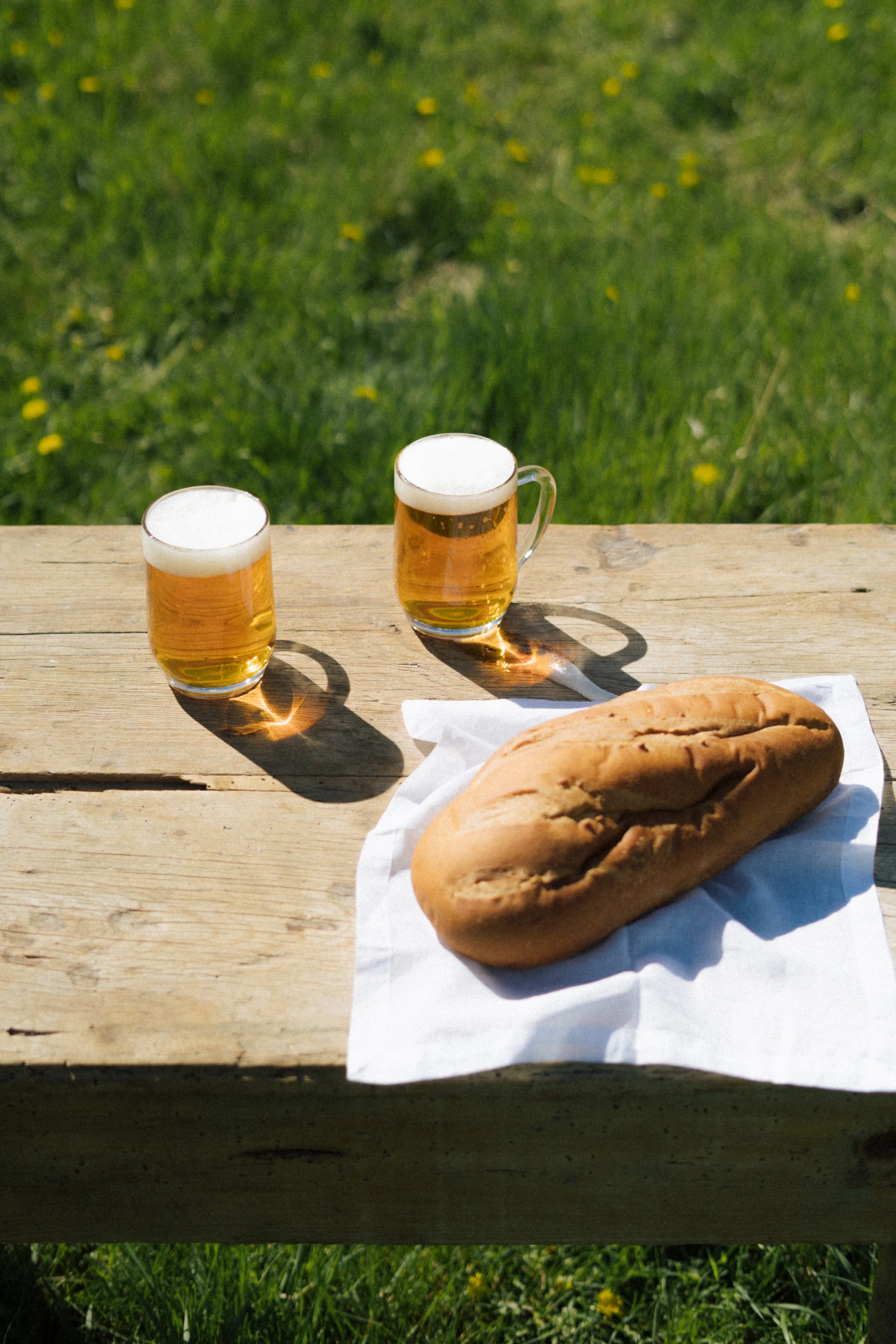 Two glasses of Bread Water beer and a baguette sandwich on a wooden table outdoors with grass and yellow flowers in the background.