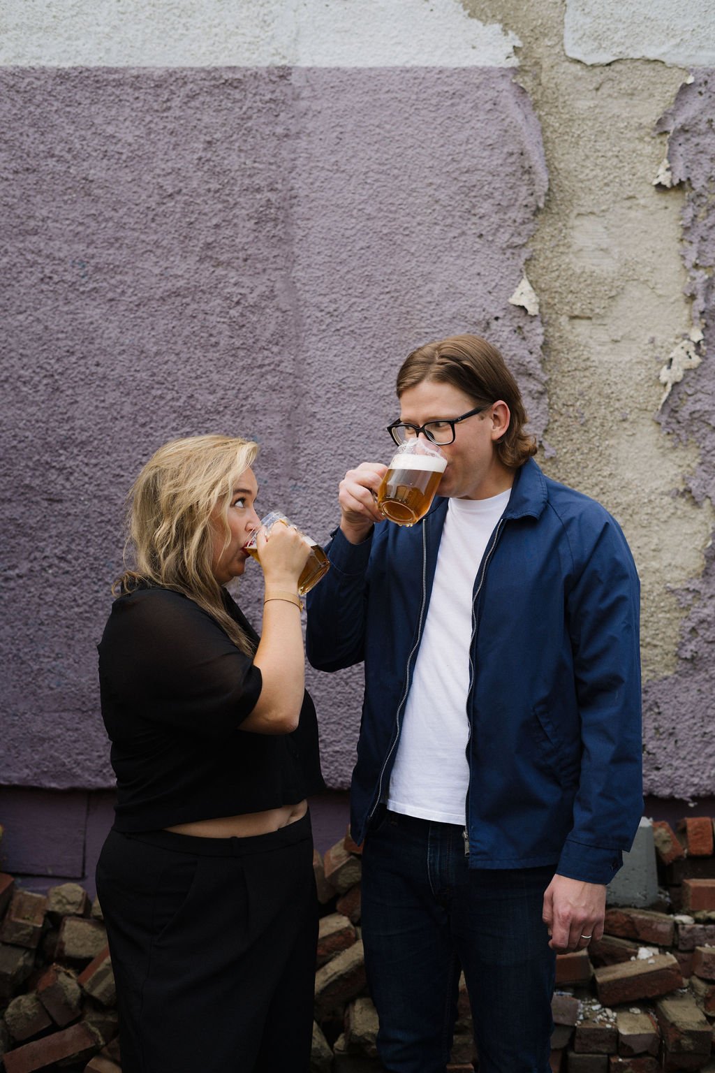 Blake and Alexa, founders of Bread Water Beer Company drinking beer together outdoors in front of a textured wall with peeling paint and bricks on the ground.