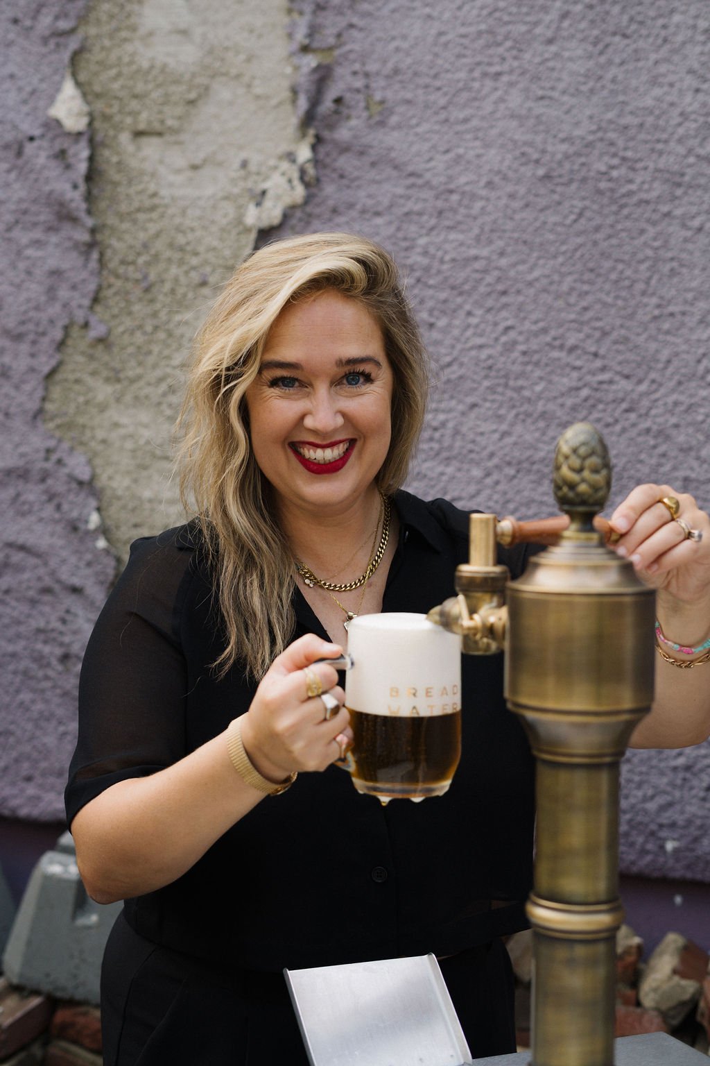 Alexa from Bread Water Beer Company, smiling, holding a mug of beer in one hand and a brass beer tap handle in the other, standing in front of a purple wall with peeling paint.