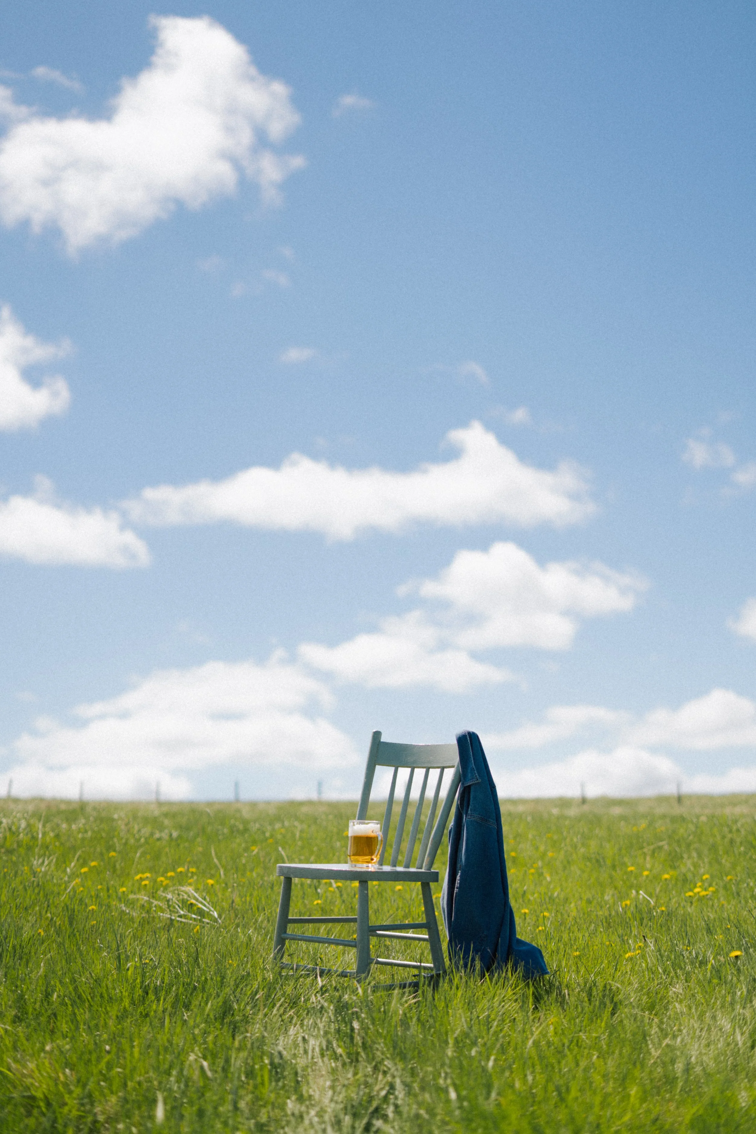 A chair with a towel draped over the backrest and a glass of Bread Water beer on the seat, set in a grassy field under a partly cloudy sky.
