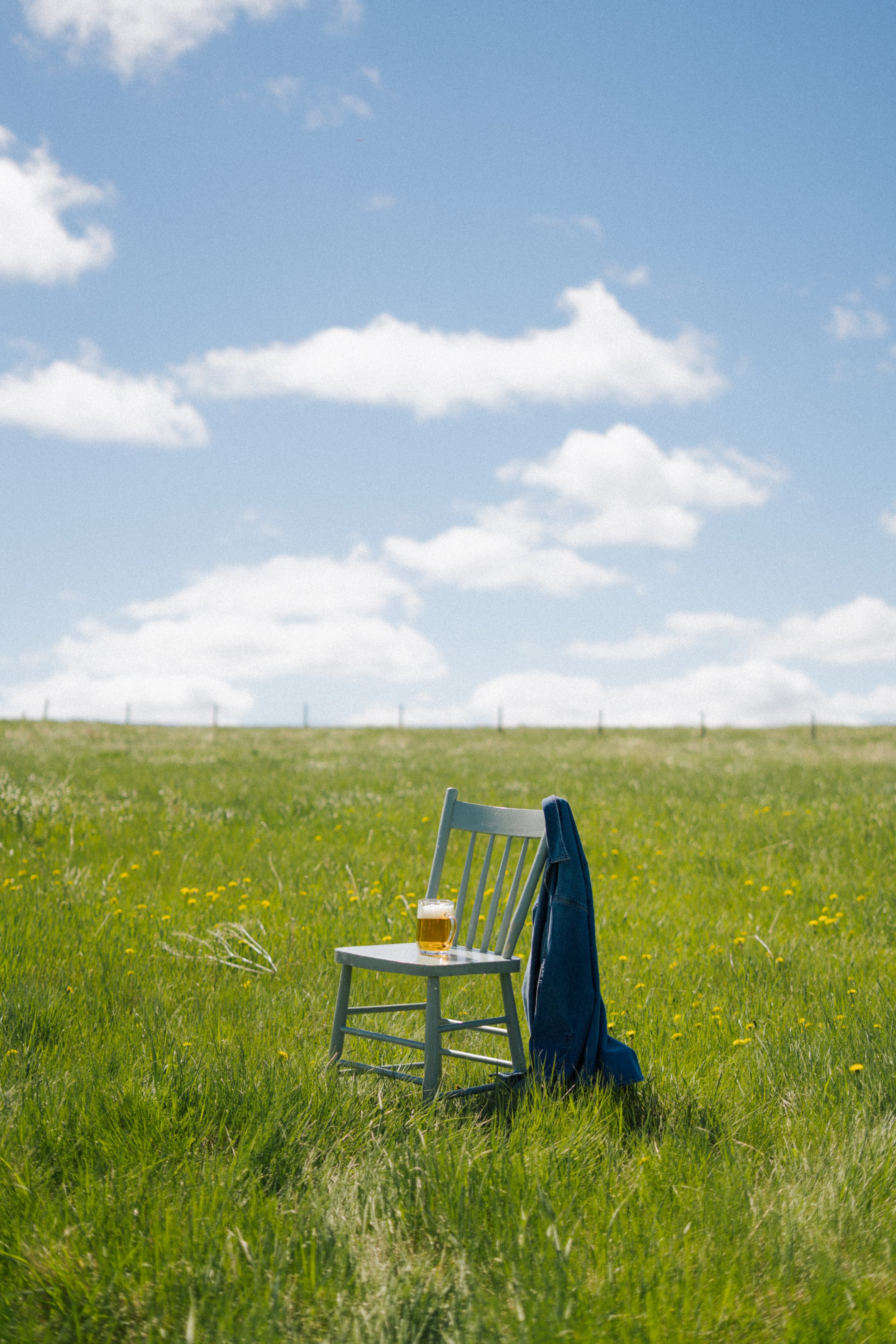 A chair with a glass of Bread Water beer on it in a grassy field with yellow flowers, a blue jacket draped over the back of the chair, and a blue jean jacket hanging on the side of the chair, under a partly cloudy sky.