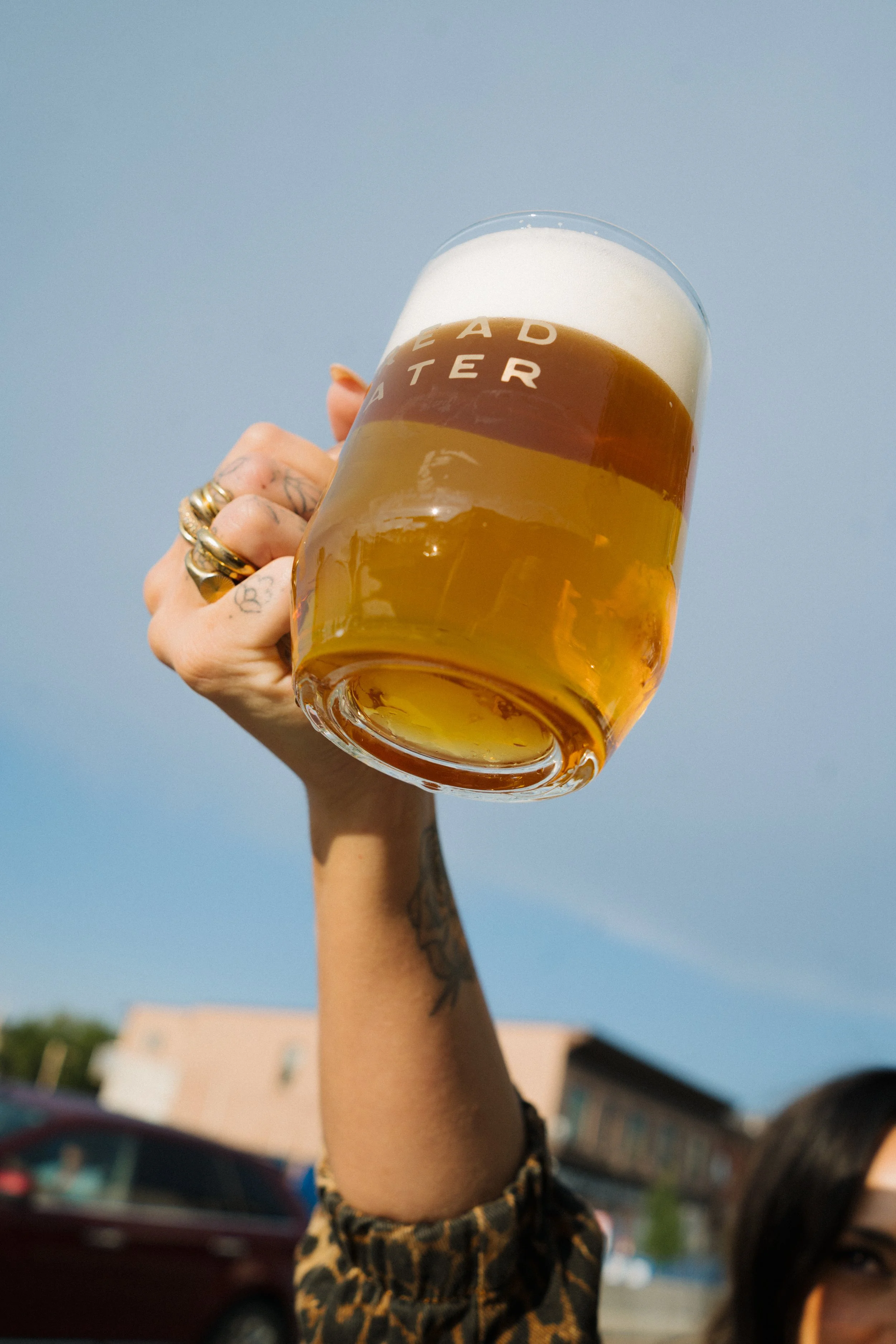 A hand with multiple rings on fingers and tattoos on wrist holding a glass of Bread Water beer with foam at the top against a blue sky.