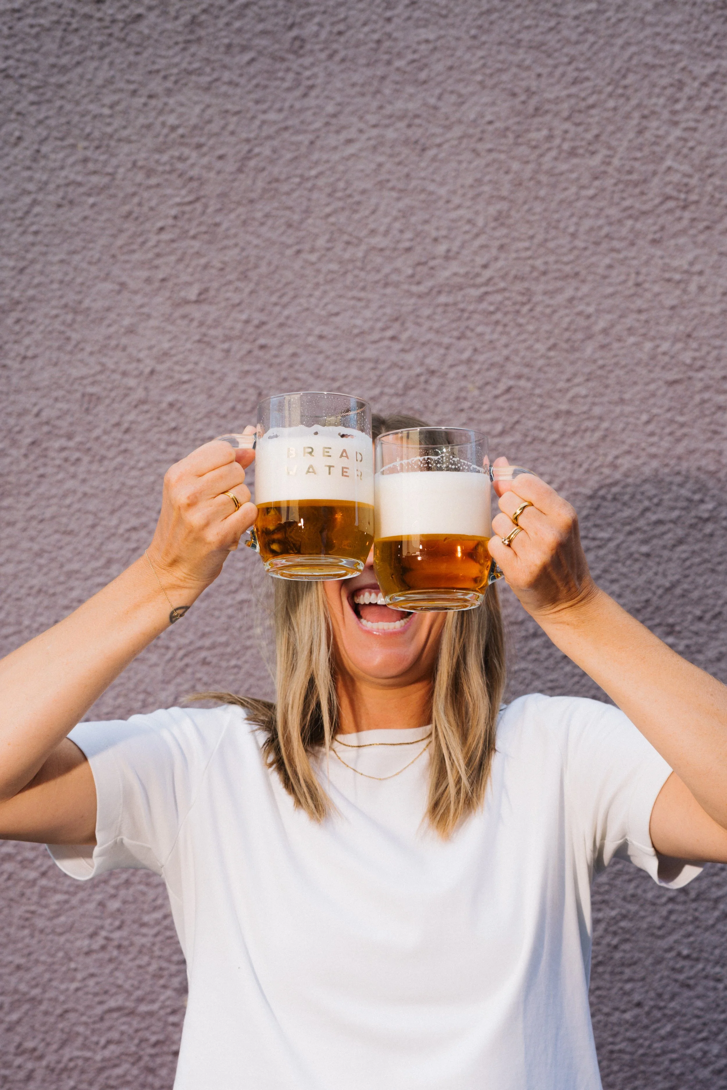 A woman holding two Bread Water beer mugs with foam on top, smiling, against a textured background.