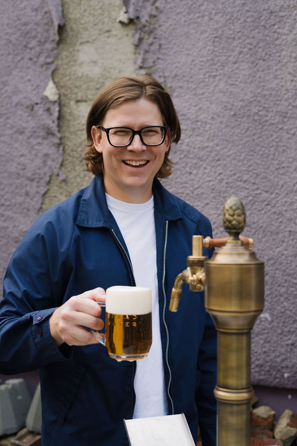 Blake from Bread Water Beer Company, wearing a navy jacket and white shirt, holding a mug of Bread Water , standing in front of a stone wall near a brass beer tap.