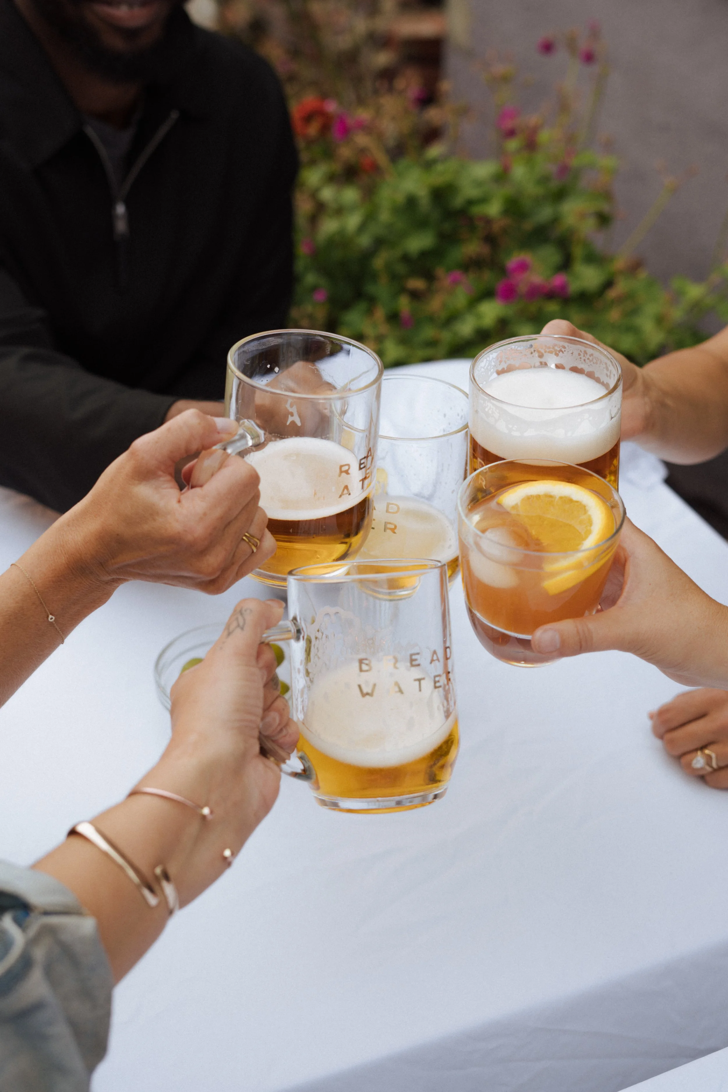 Group of people clinking glasses of Bread Water beer and cocktails with lemon slices, celebrating outdoors against a background of colorful flowers and greenery.