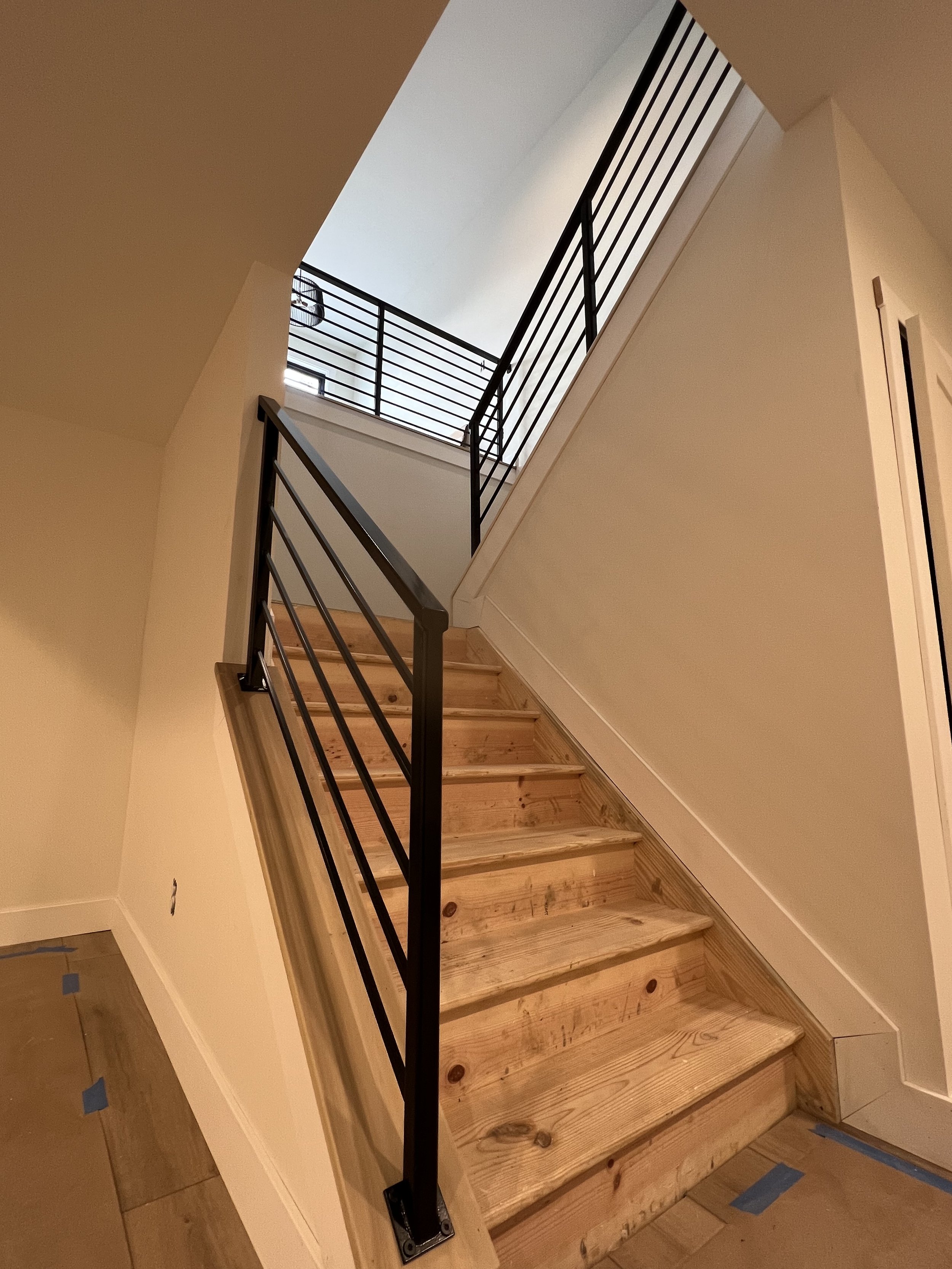 Interior view of a staircase with wooden steps and a black metal railing, leading to an upper level with a ceiling window and white walls.