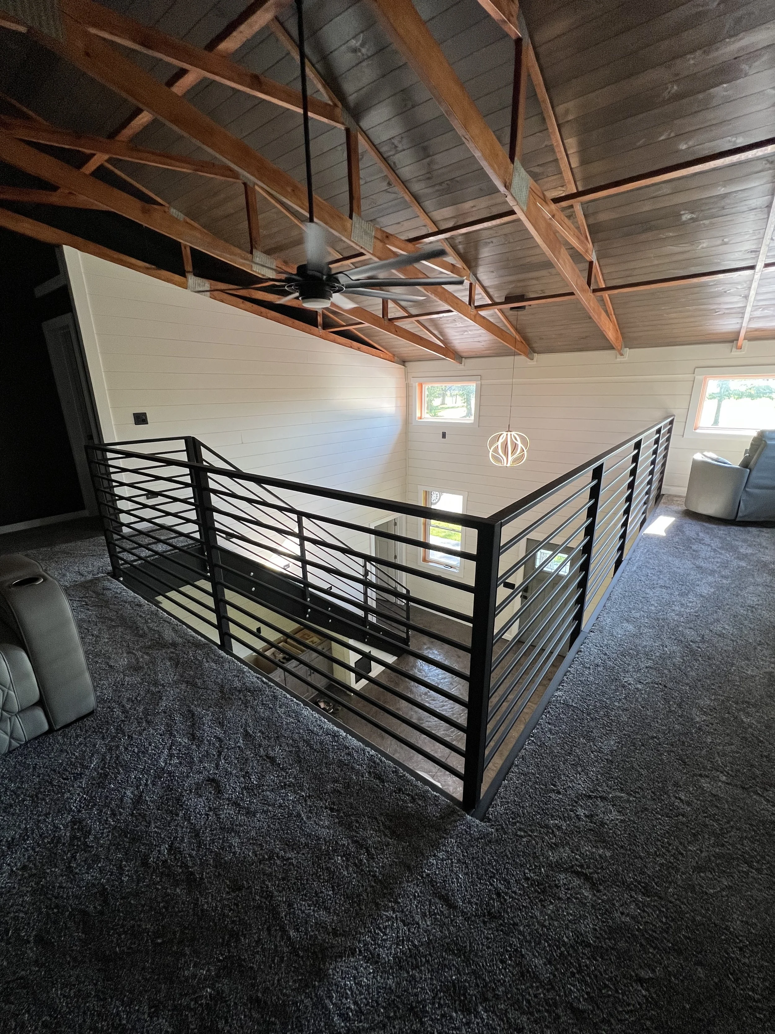 Interior view of a house showcasing a second-floor balcony with a black metal railing, a ceiling fan, and a modern light fixture hanging from the ceiling. The ceiling features exposed wooden beams, and the walls are painted white, with several windows allowing sunlight to enter.