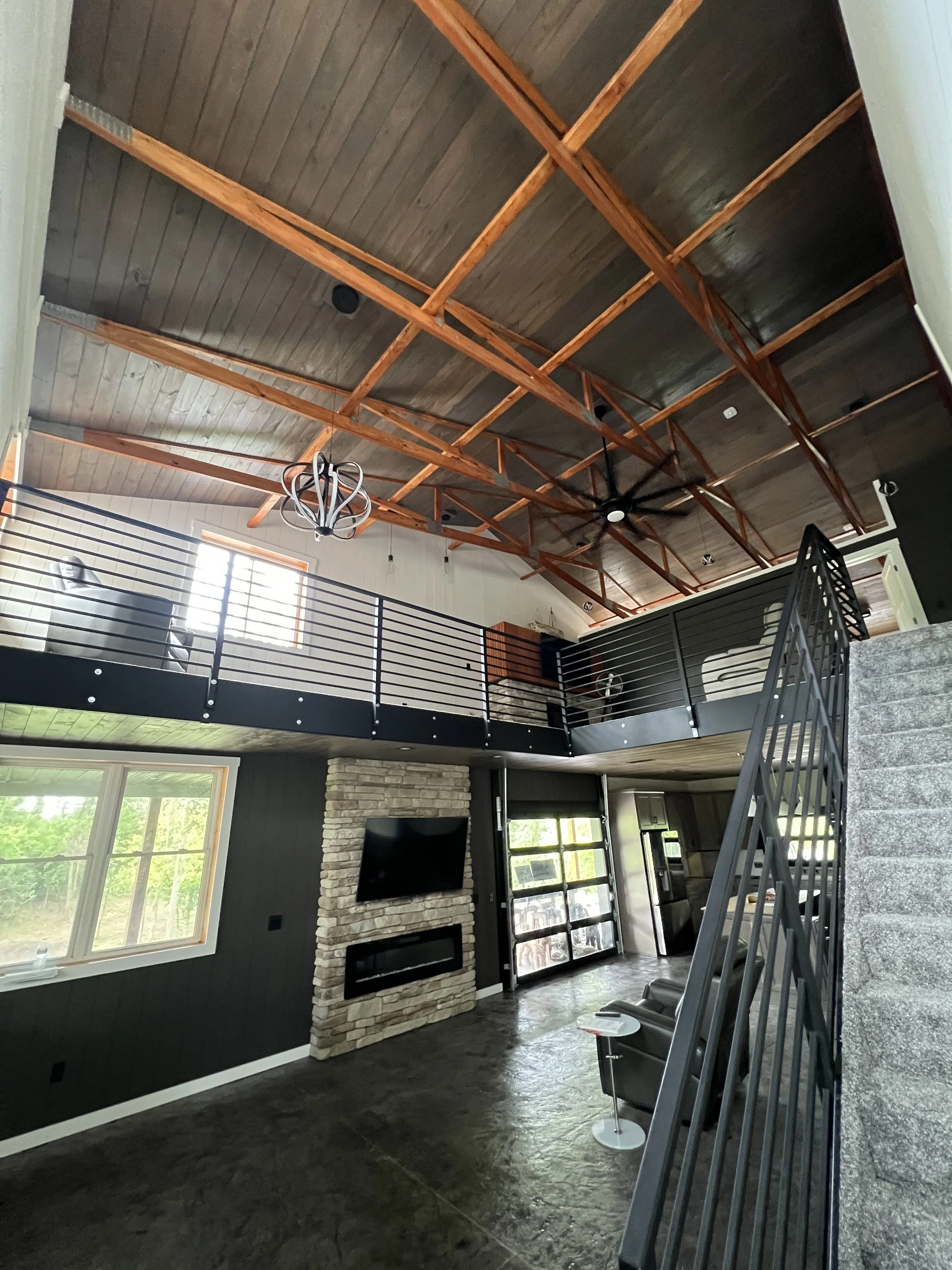 View of a modern living room with a high wooden ceiling supported by exposed beams, a stone fireplace with a wall-mounted TV, large windows letting in natural light, and a black metal railing on the second floor.