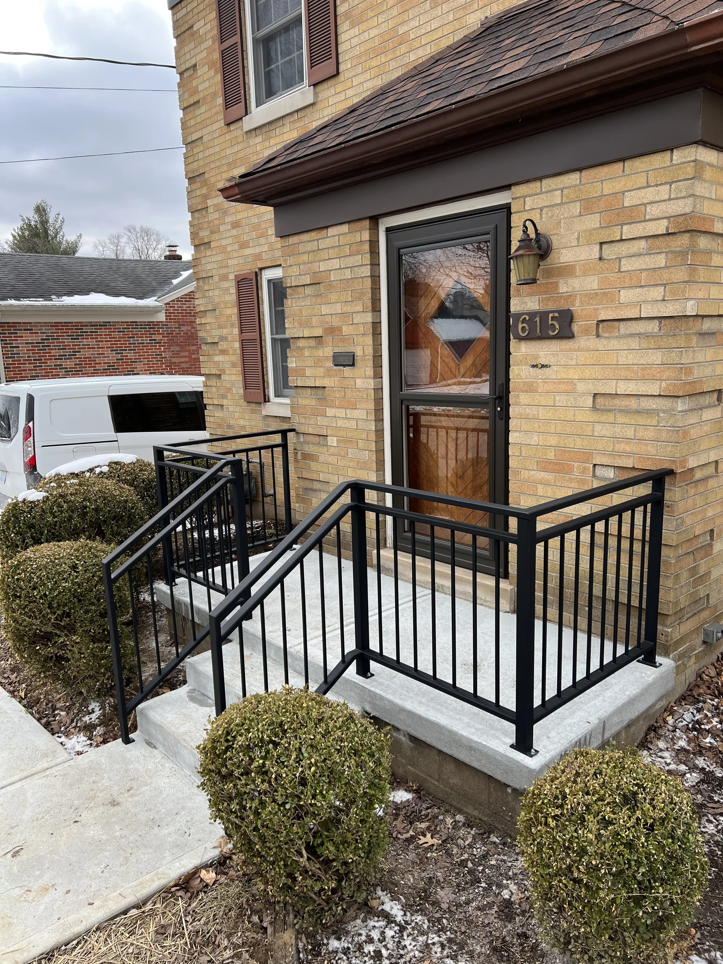 Exterior of a brick house with a black metal railing and concrete steps leading to the front door, with trimmed bushes and patches of snow on the ground.