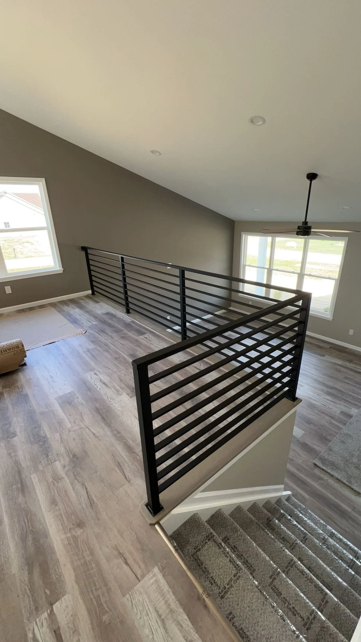 Interior view of a modern two-story home, showing an open living area with large windows, wooden flooring, a staircase with carpeted steps, and a black metal railing along the edge of the upper floor.
