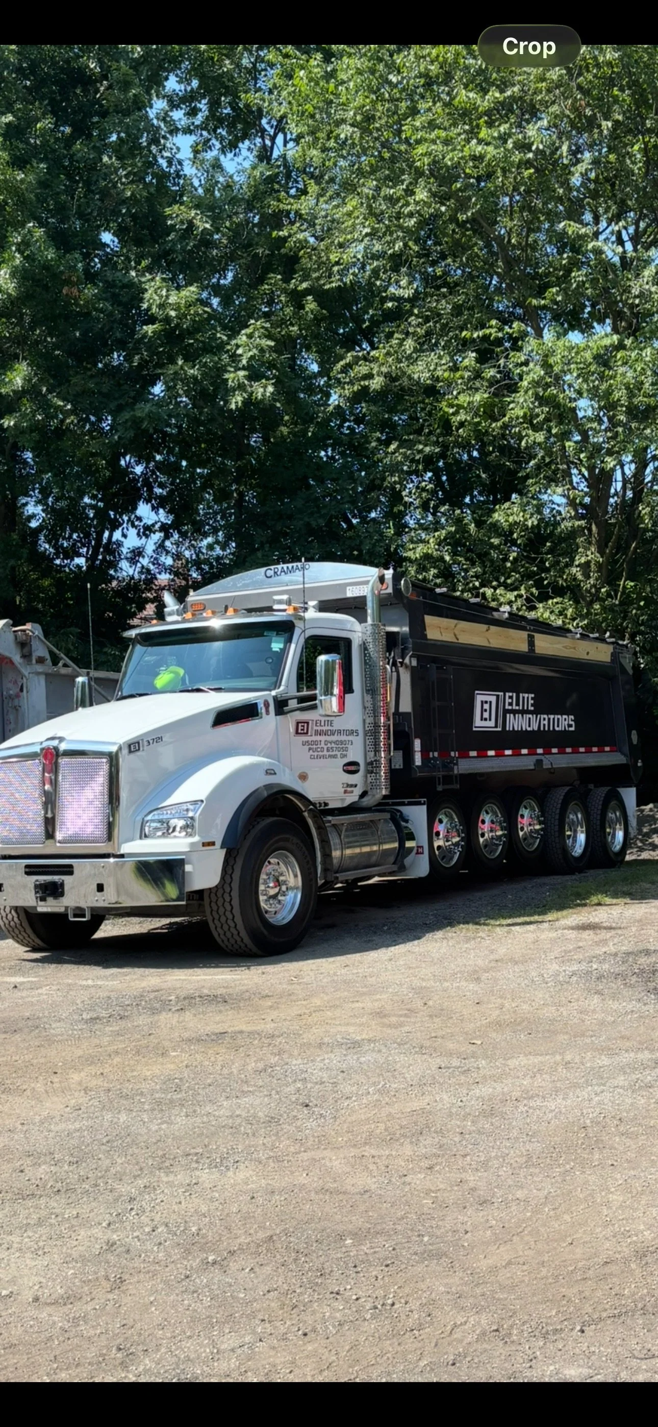 A white and black dump truck with 'Elite Innovators' logo parked on a dirt lot under green trees with clear blue sky.
