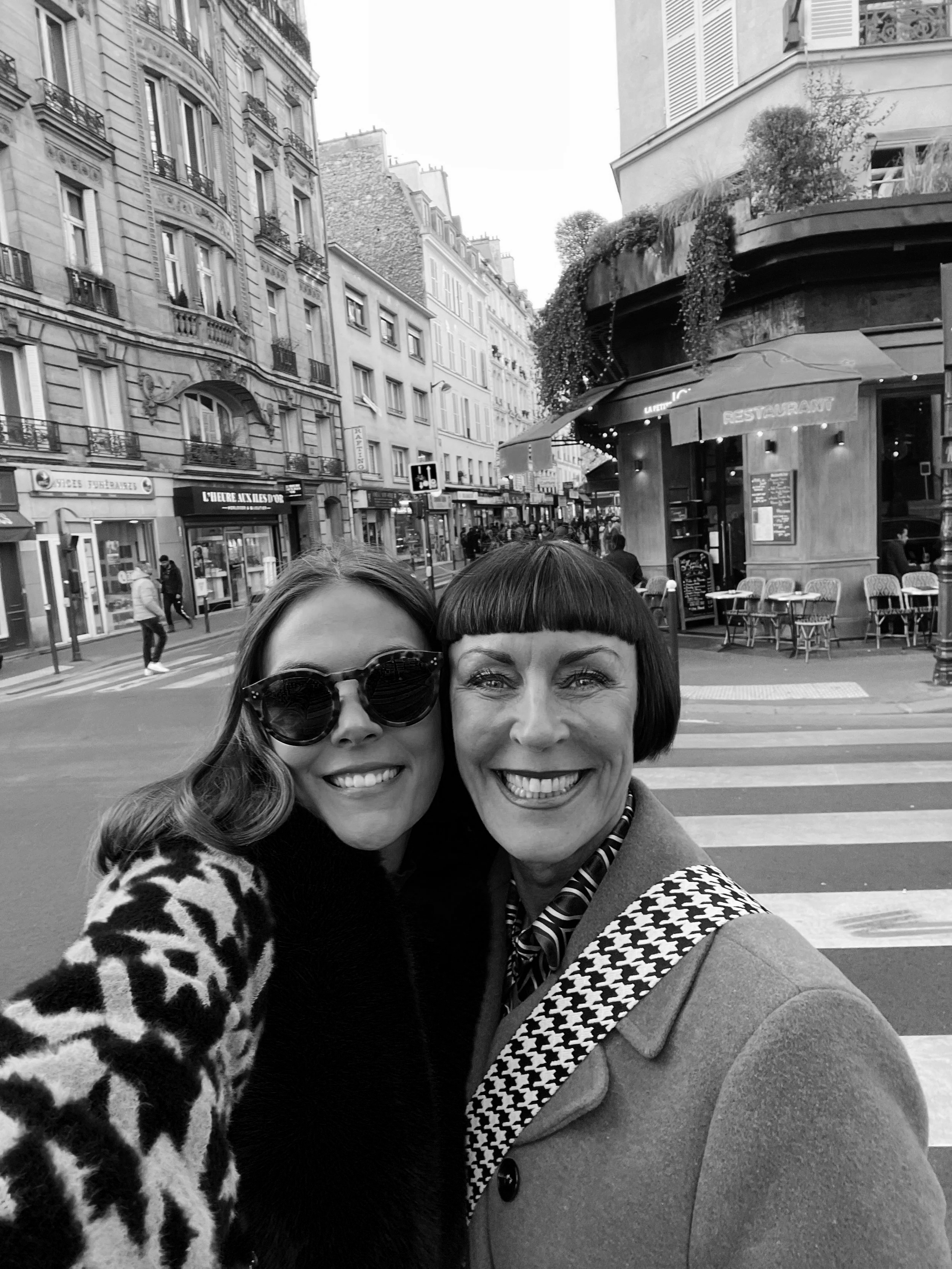 Two women taking a selfie on a city street, with buildings, storefronts, and pedestrians in the background.