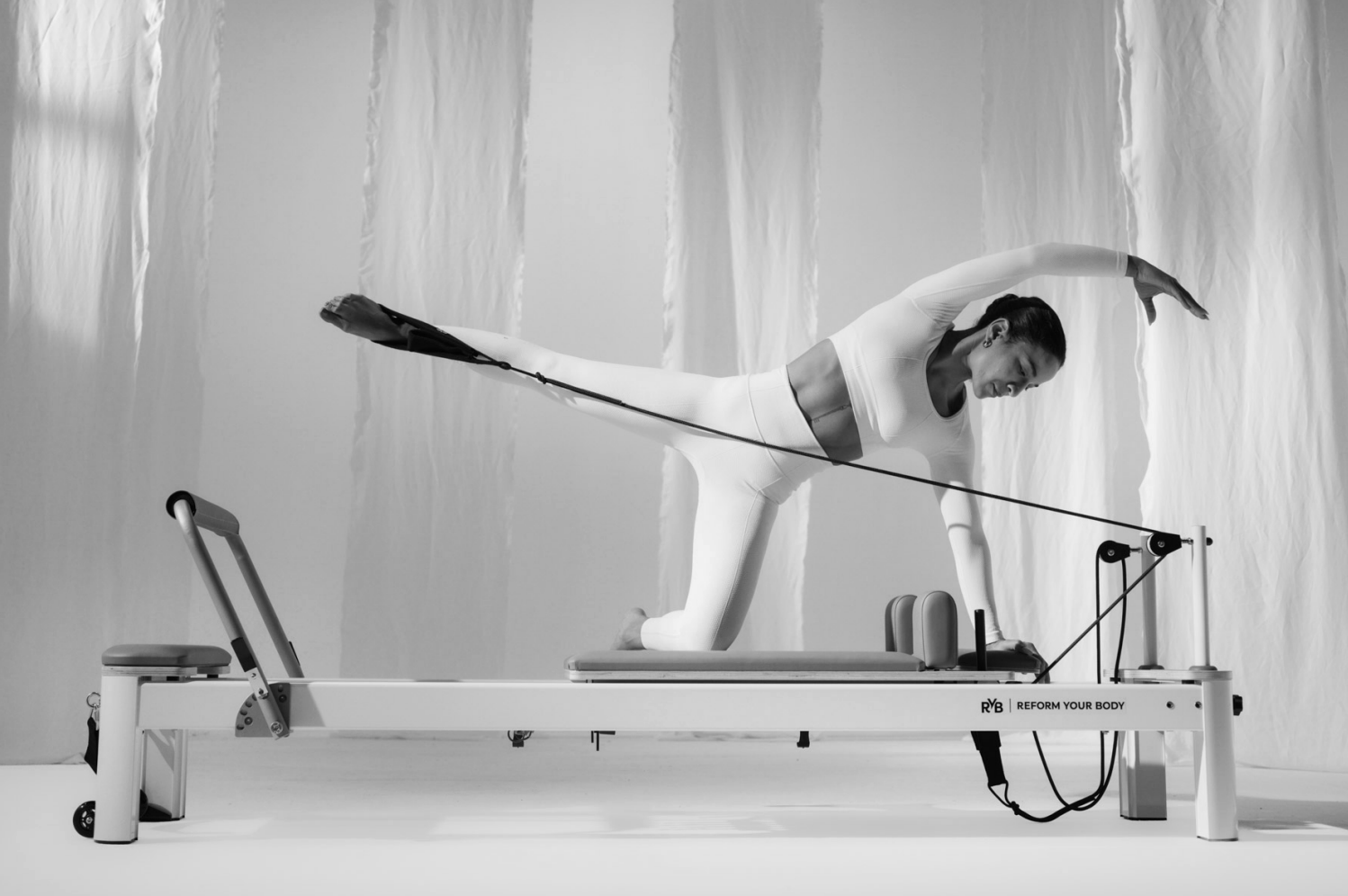 A woman in athletic clothing performing a stretching exercise on a reformer machine in a studio.