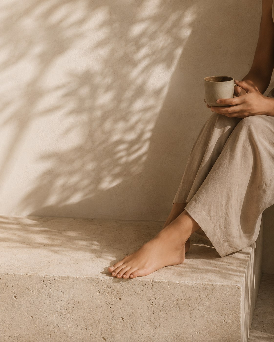 Person sitting on a beige stone ledge holding a ceramic cup, with sunlight casting textured shadows on the wall.