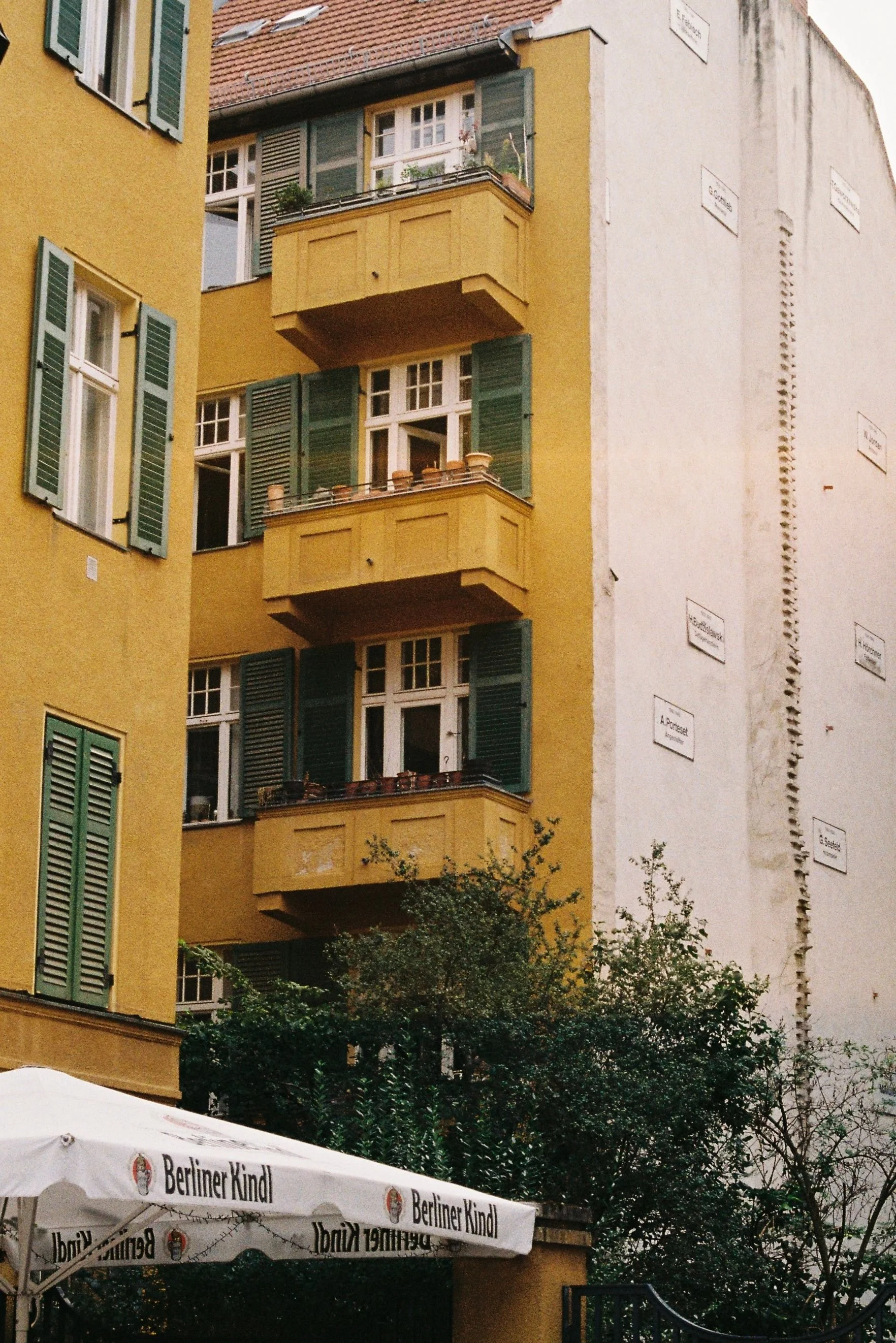 Yellow building with green shutters and balconies, some with plants, and a white side wall with street signs, below which is an outdoor seating area with a Berliner Kindl branded umbrella surrounded by trees.