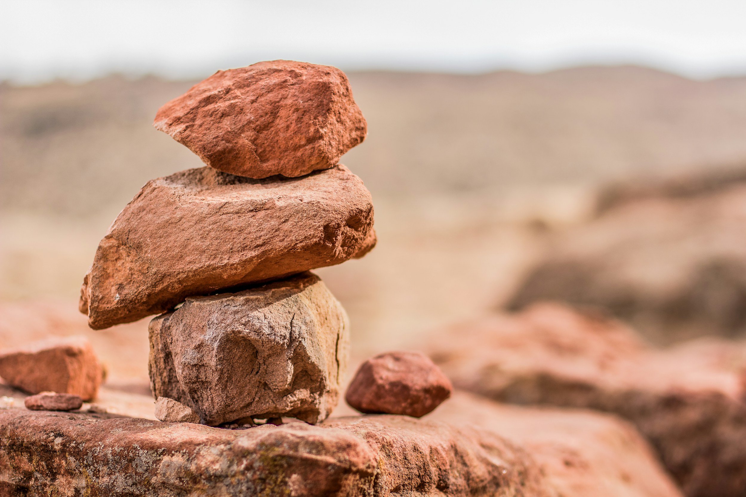 A stack of four reddish-brown rocks balanced on top of each other on a sandy surface, with a blurred background of more rocks and sand under an overcast sky.