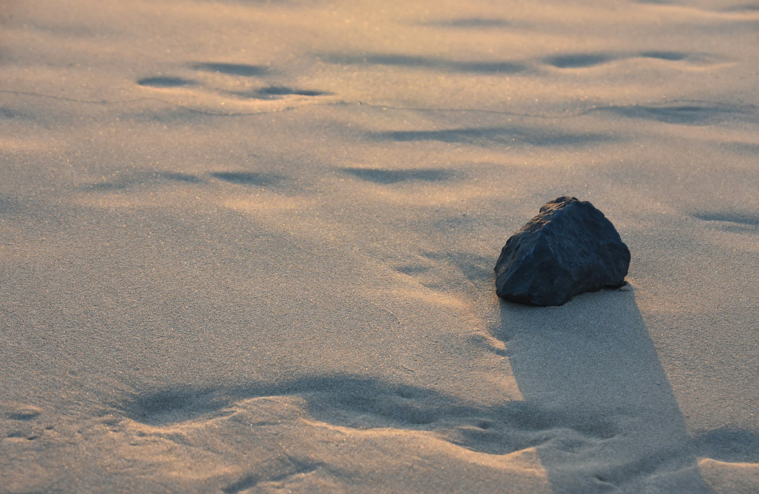 A single dark rock on a sandy beach with gentle shadows cast by the setting or rising sun.