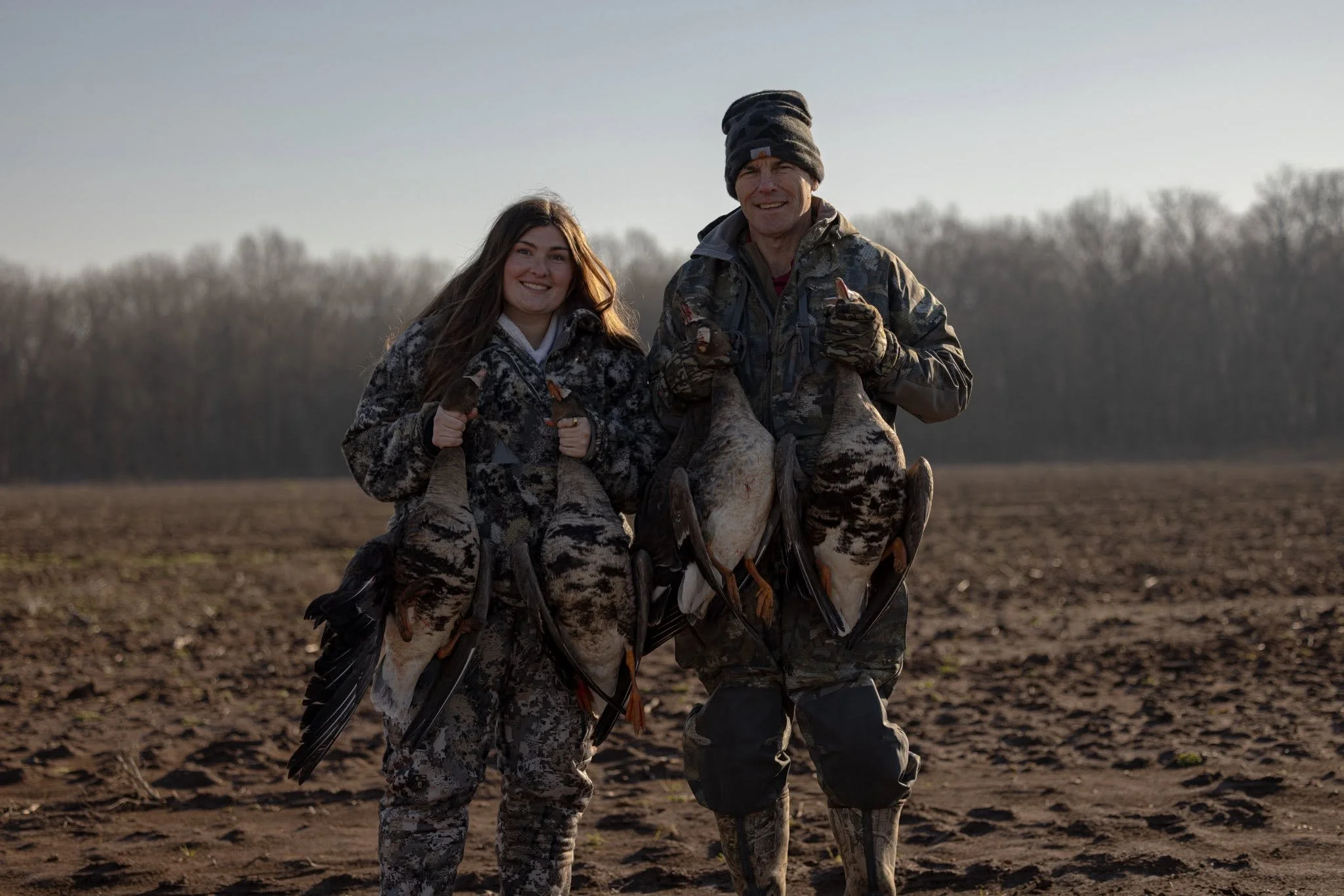 A young woman and an older man in camouflage hunting gear posing outdoors with a dead goose. The woman is smiling and holding the goose, while the man is pointing at it and smiling.
