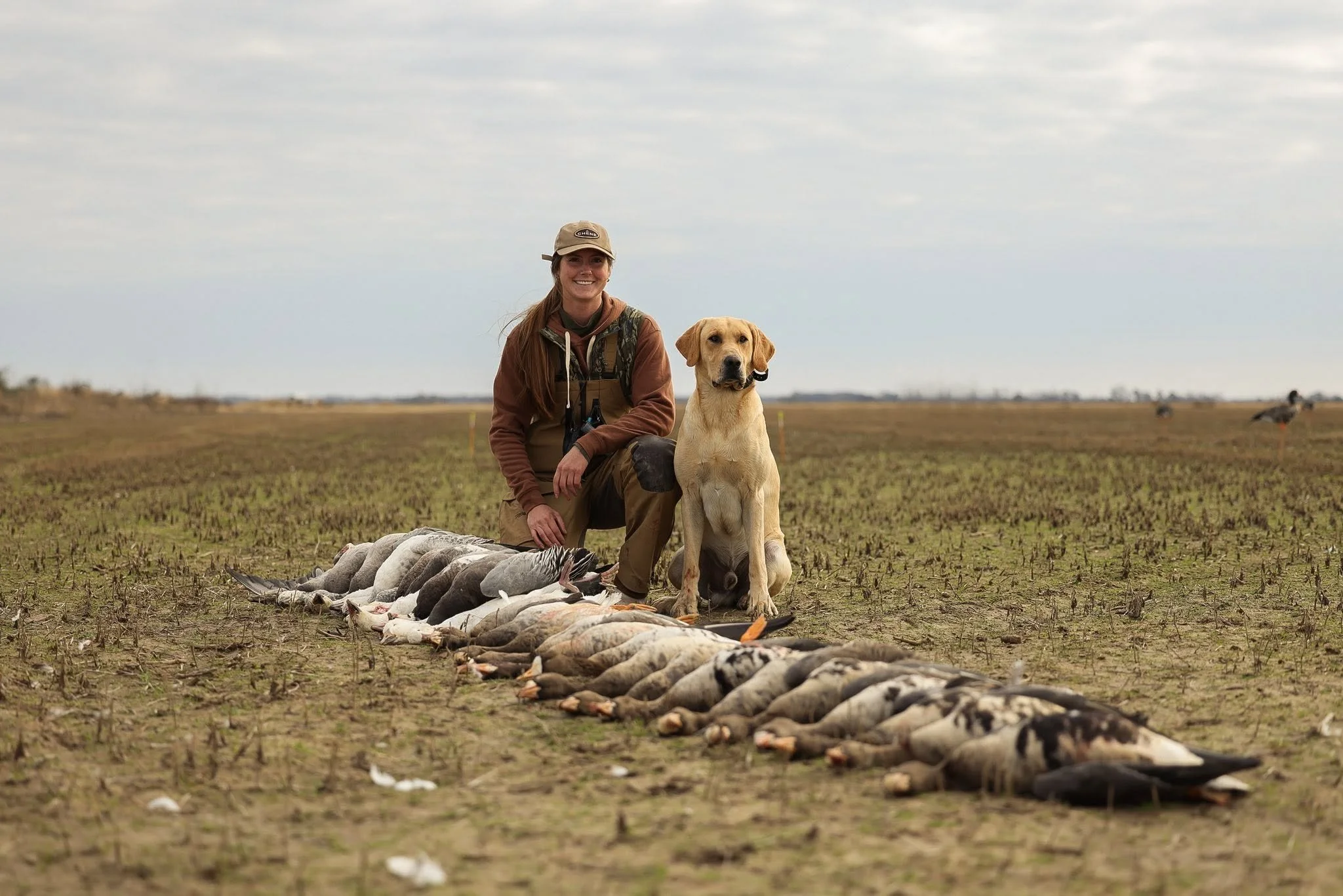 A woman with a dog kneeling next to a line of dead geese on a field during daytime. The woman is smiling and wearing outdoor gear, and the sky is cloudy.