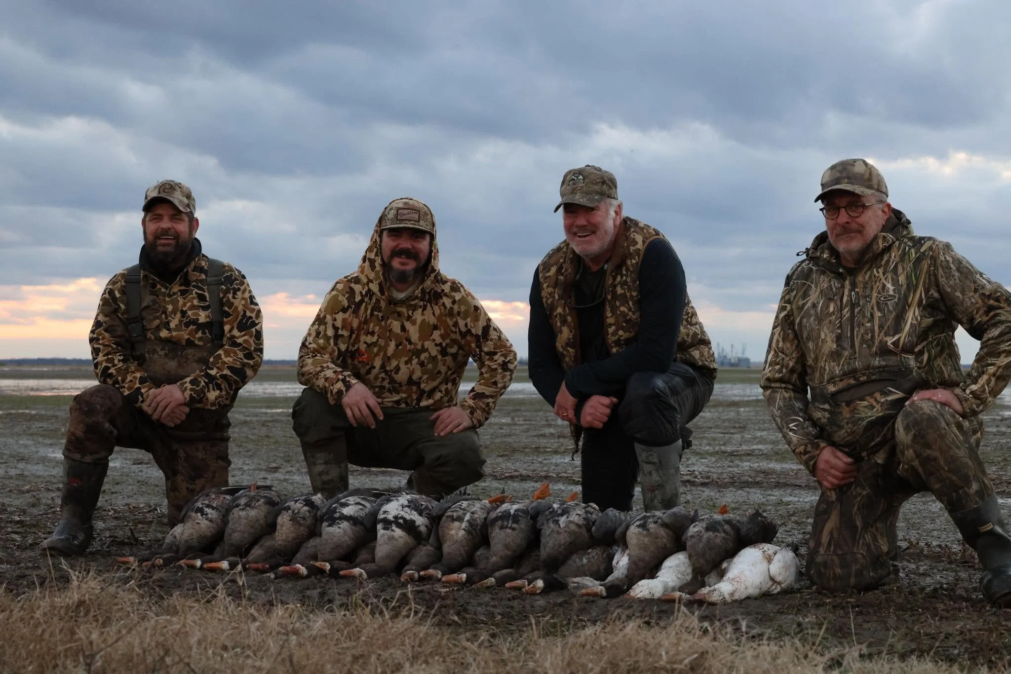 Four men dressed in camouflage outdoor clothing and hats, kneeling or squatting outdoors on a muddy terrain with a cloudy sky in the background, posing in front of a row of geese after a guided specklebelly goose hunt.