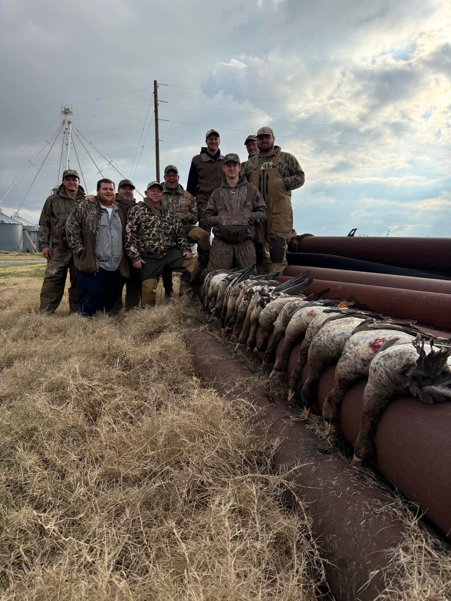 Group of people posing outdoors during daytime, dressed in hunting or outdoor gear, with geese laid out in front of them, and farm structures and power lines in the background.