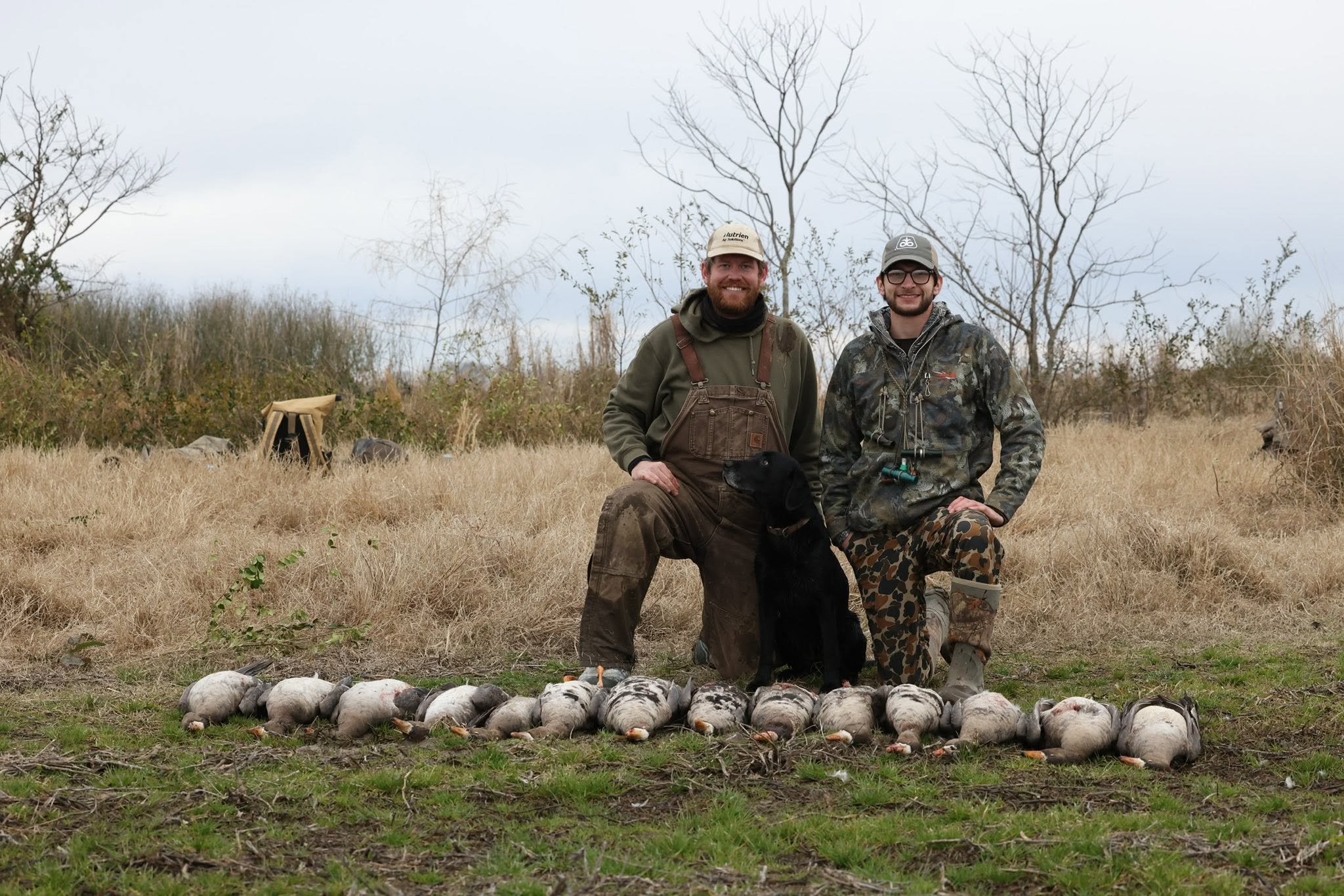 Two men kneeling in a field of harvested rice, smiling, with a row of hunted geese laid out in front of them. Bare trees and overcast sky in the background.