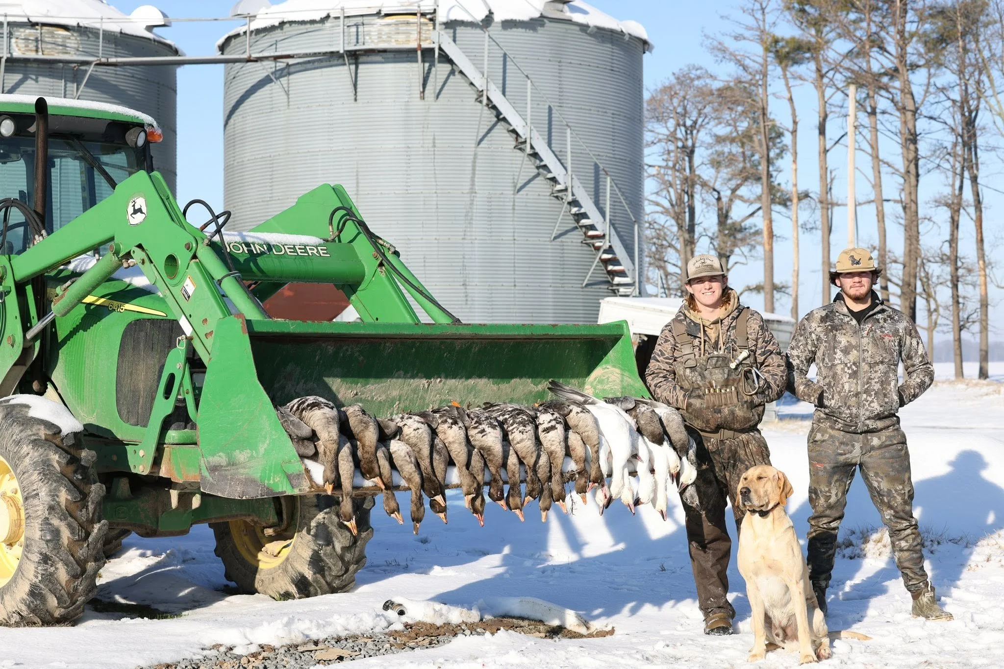 Two people dressed in camouflage standing outdoors in winter, holding bags of hunted game, with a golden retriever in front and a green John Deere tractor with a load of hunted geese on it in the snow, and large grain silos in the background.