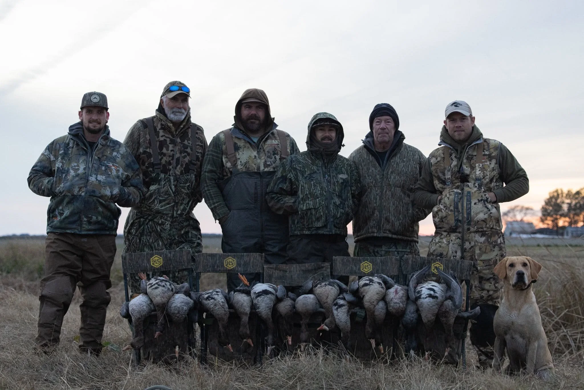 A group of six men in camouflage and outdoor clothing standing outdoors during dusk, with a dog, a row of hunted game birds, and sporting equipment in front of them, likely after a hunting trip.