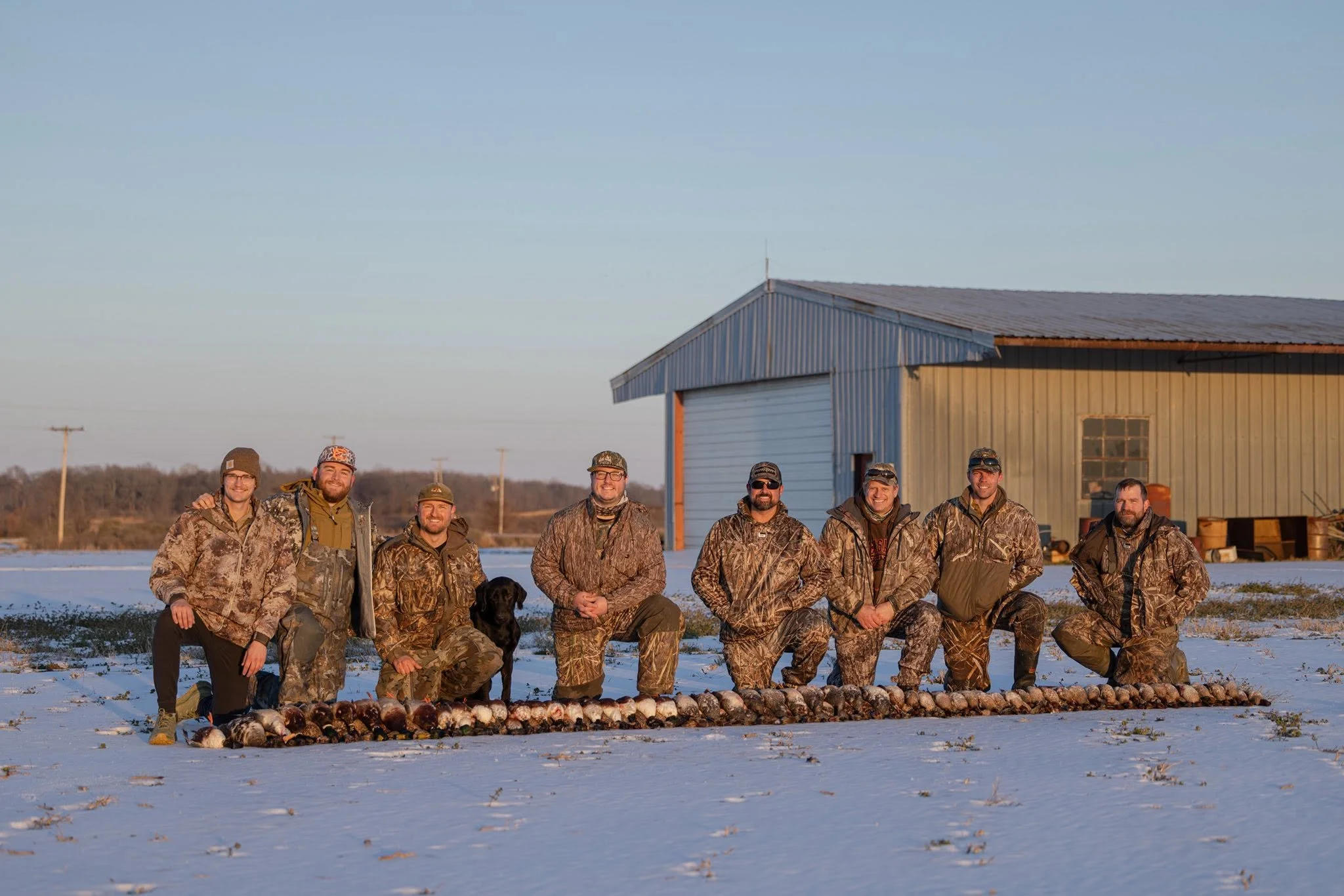 Group of eight people dressed in camouflage clothing, kneeling and standing on snow-covered ground, with a row of dead birds laid out in front of them, and a black dog among the group, in front of a metal building and utility poles at sunset or sunri