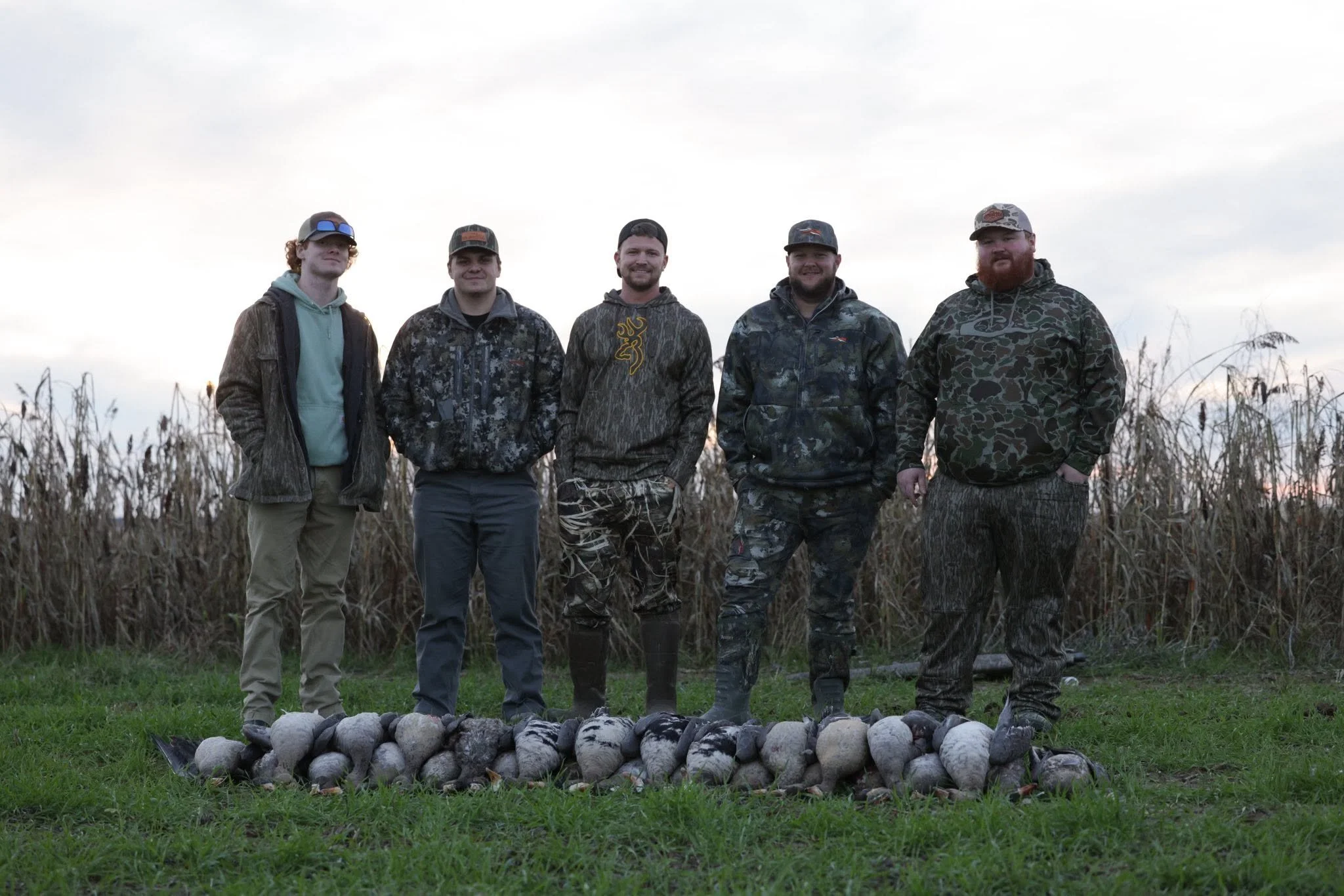 Five men standing outdoors in a line, dressed in camouflage and outdoor clothing, with a pile of geese front of them