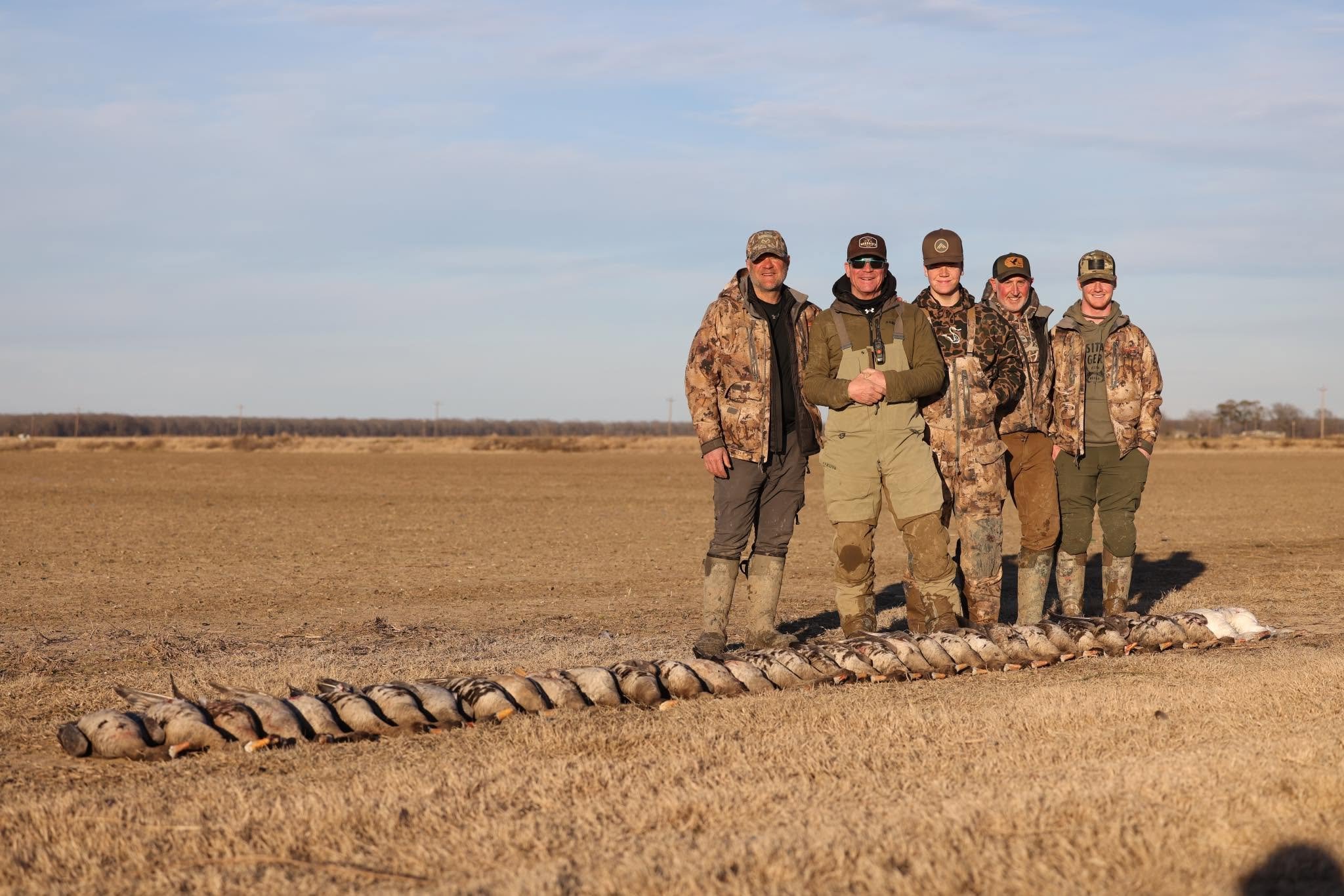 Five men in camouflage and outdoor gear standing outdoors with a line of dead geese laid out on the ground in front of them.