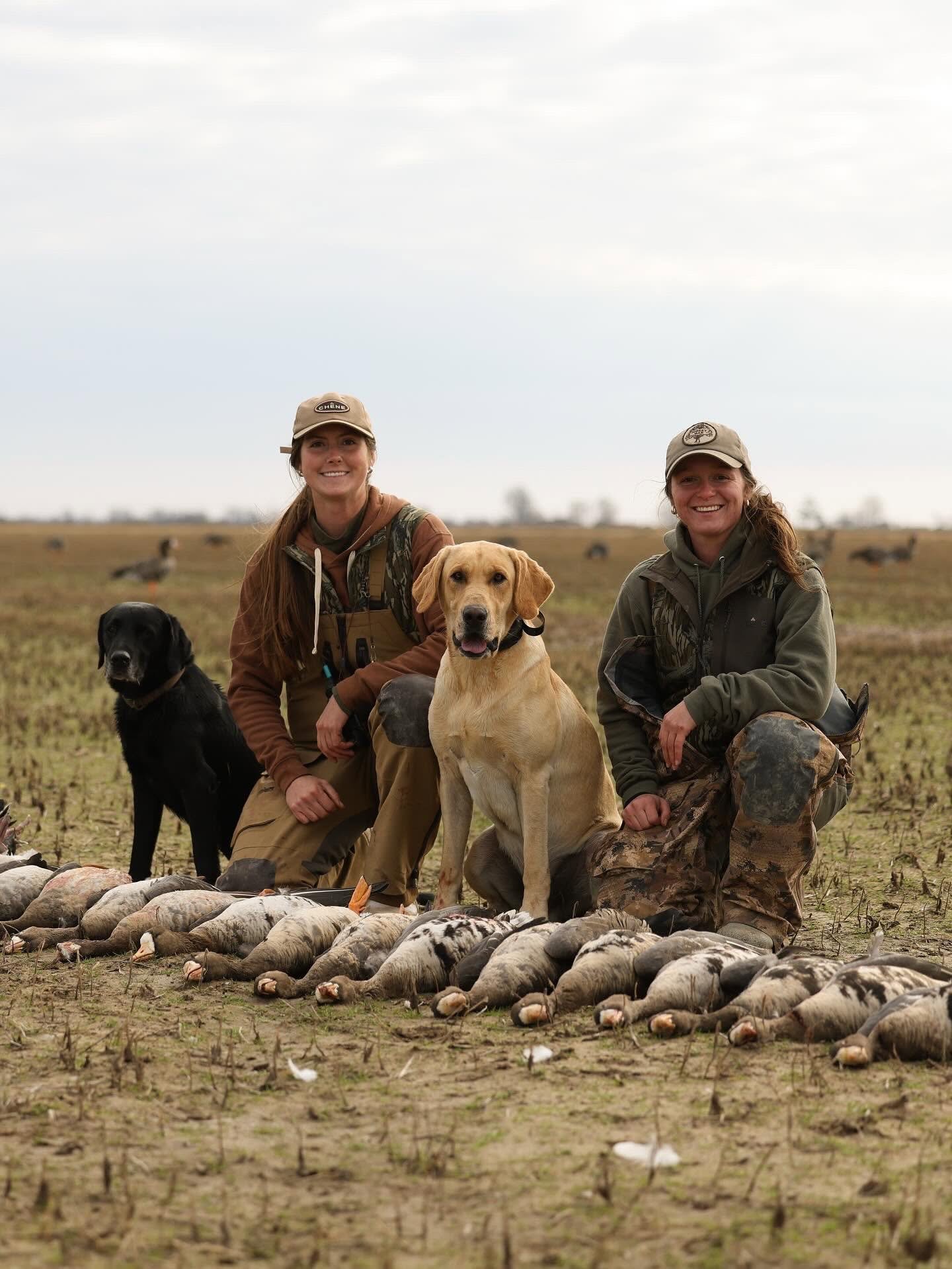 Two women dressed in outdoor hunting gear with dogs, kneeling and smiling, surrounded by freshly hunted geese on an open field, with a flock of waterfowl in the background.