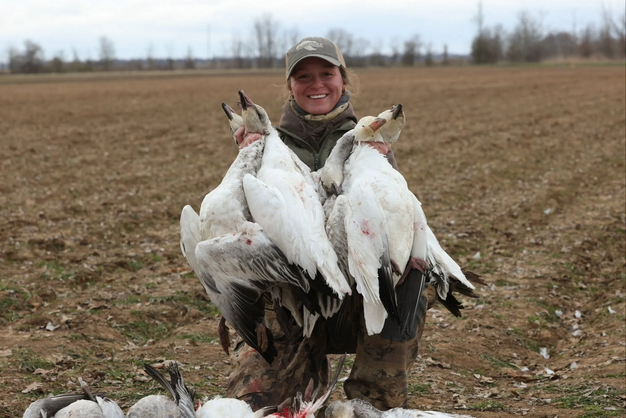 A woman in outdoor clothing holding several drowned geese, standing on a field with a cloudy sky in the background.