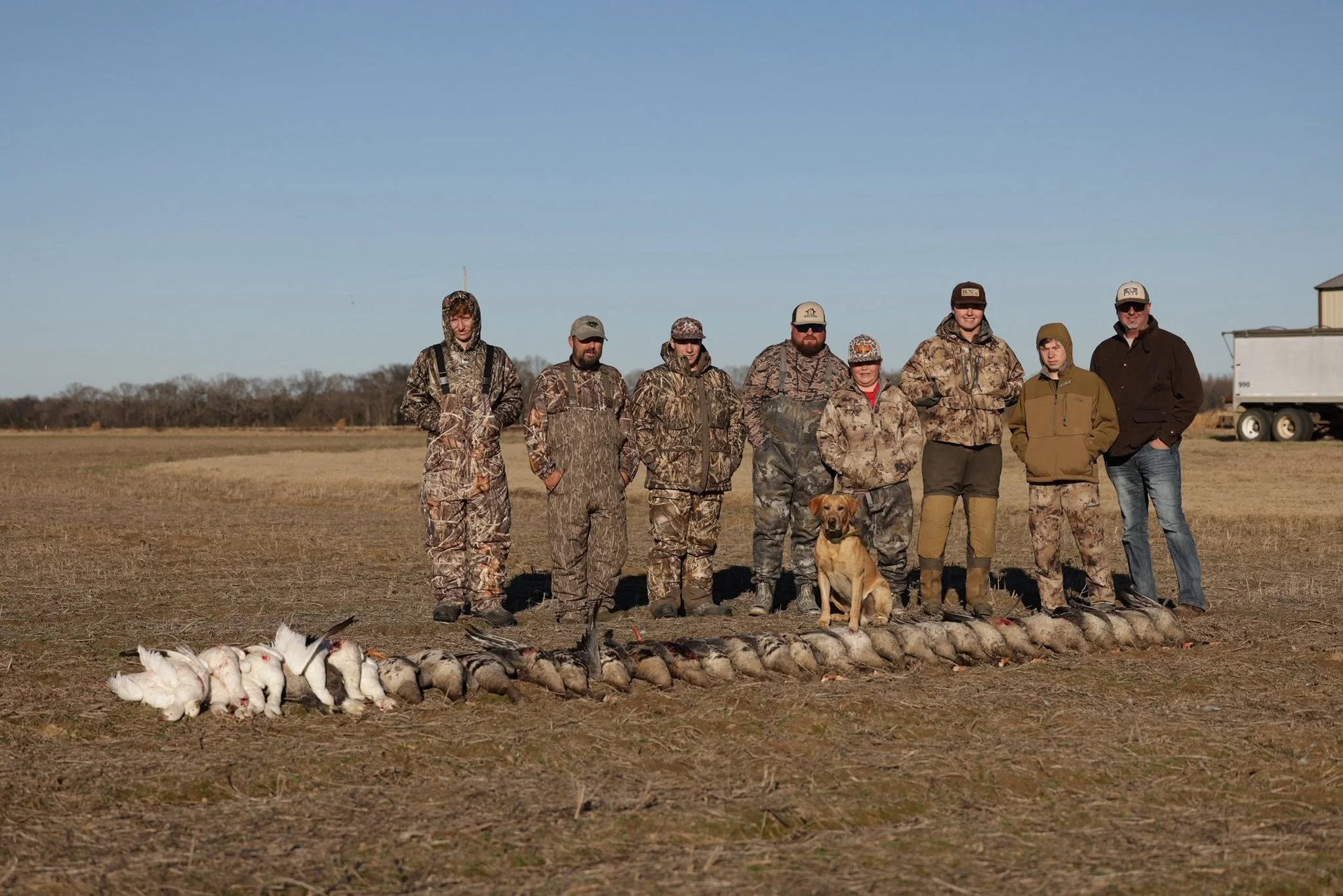 Group of eight people dressed in camouflage and outdoor clothing standing in a field with birds and a hunting dog in front of them, under a clear blue sky.