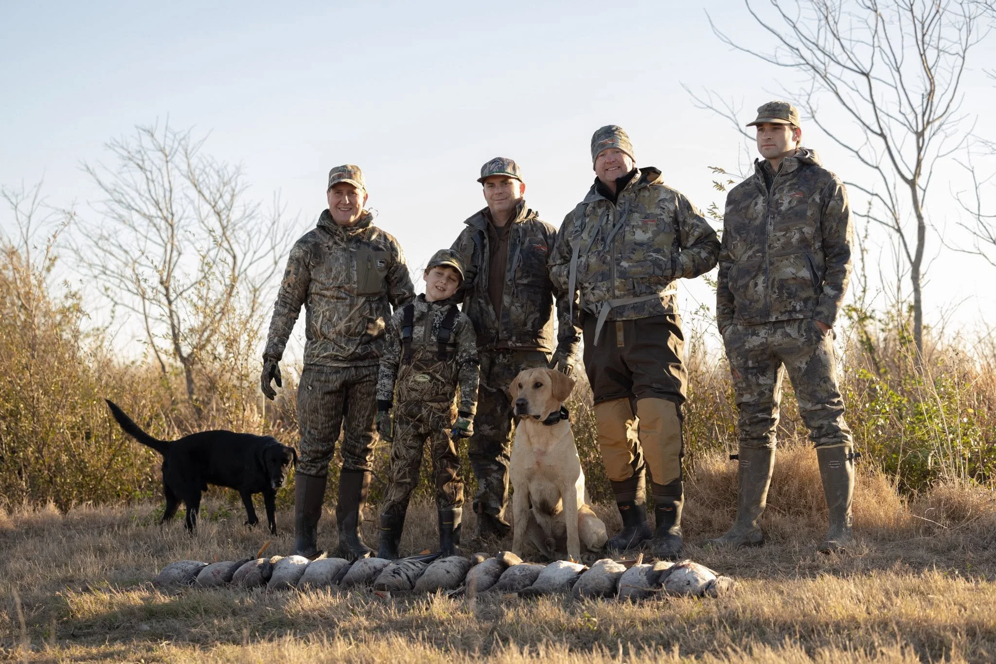 A group of four adults and a child in camouflage hunting jackets and hats, posing outdoors with two hunting dogs, a black and a yellow Labrador, in front of a row of harvested game animals, with leafless trees and a clear sky in the background.
