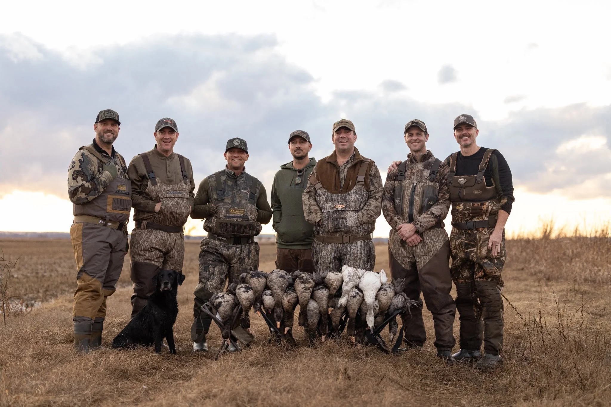 A group of seven men dressed in camouflage and outdoor gear standing outdoors in a field, smiling at the camera, with a black dog sitting nearby. 