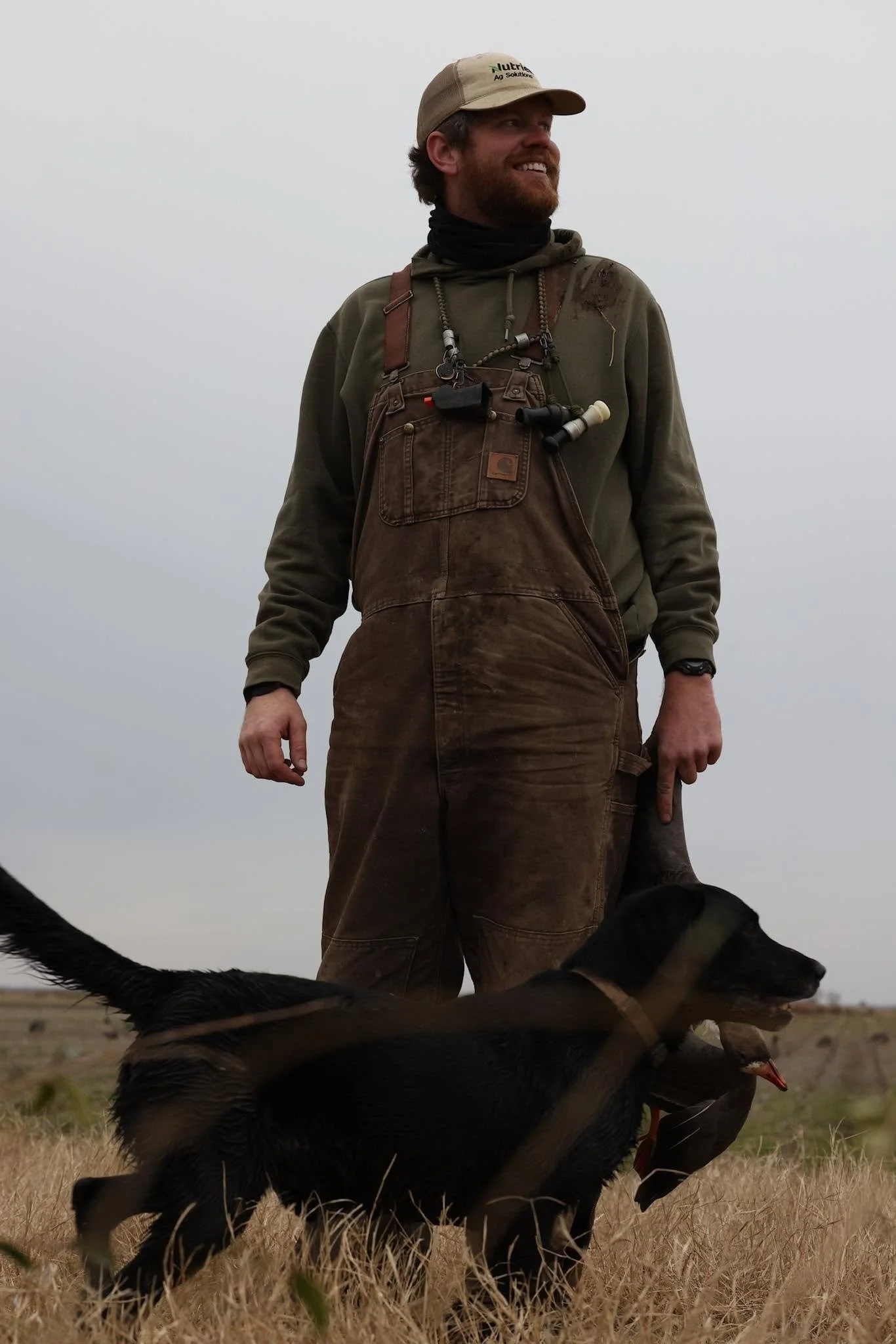 A man with a beard, smiling, wearing a beige cap, green sweatshirt, and brown overalls, outdoors in a field with dry grass