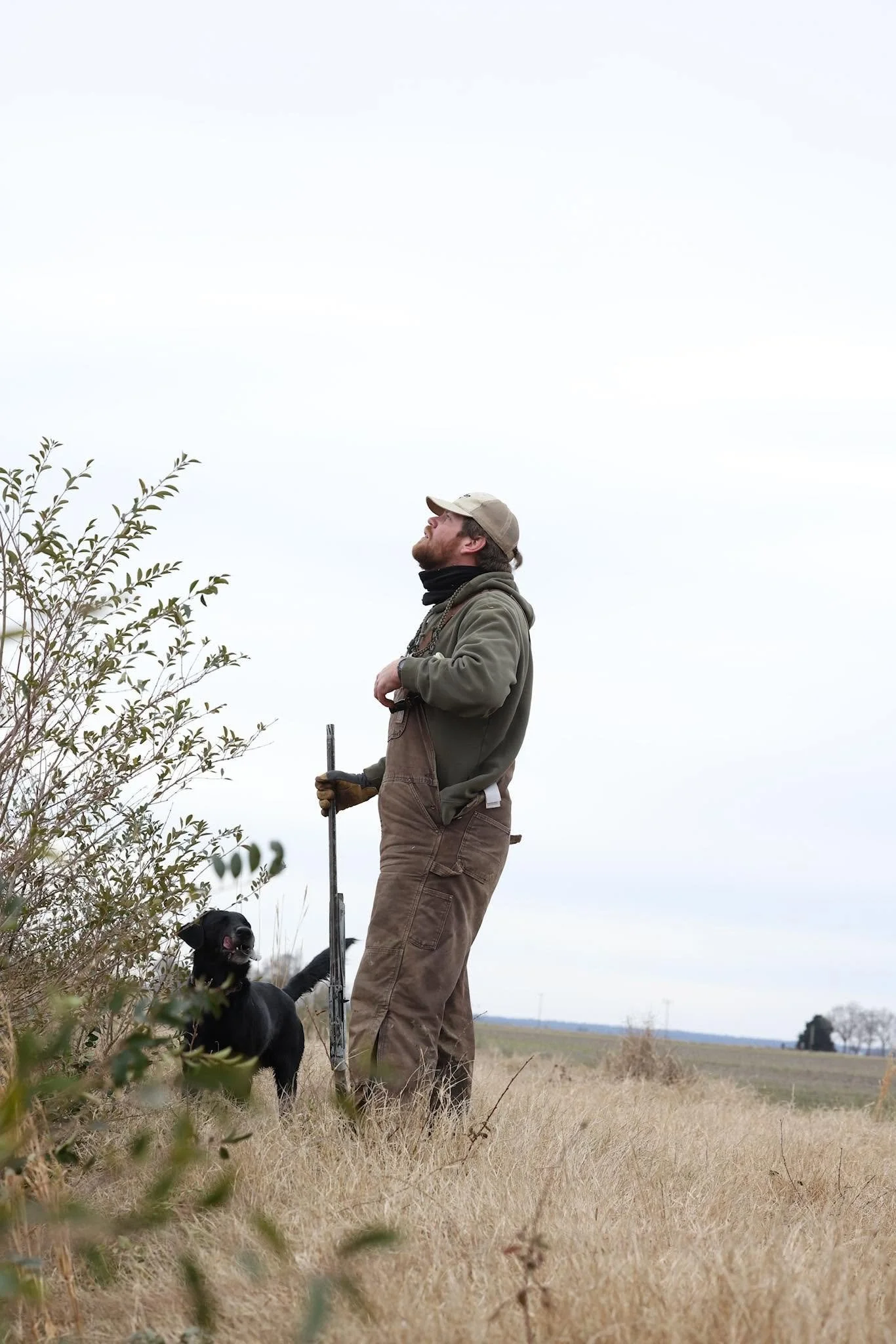 A man with a beard and tan cap standing outdoors in a field, holding a tall stick, with a black and white dog sitting nearby. 
