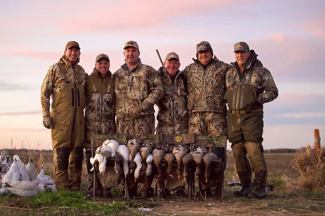 A group of six men in camouflage and outdoor gear standing outdoors at sunset, posing with a limit of geese laid out in front of them after a hunting trip.