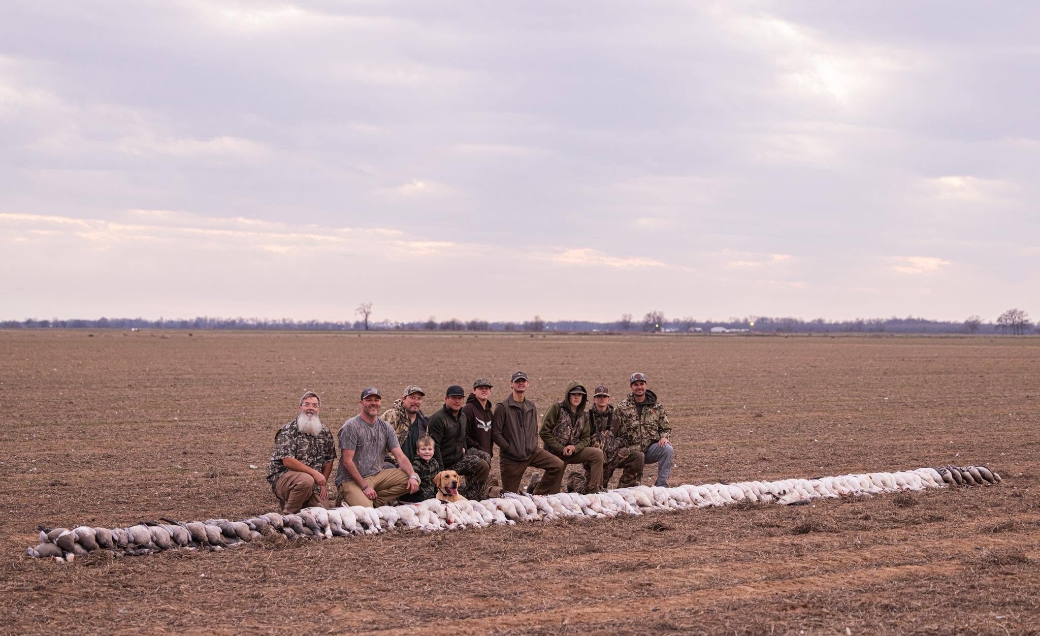 Group of people, including men, women, children, and a dog, posing outdoors on a farm field with a cloudy sky in the background.