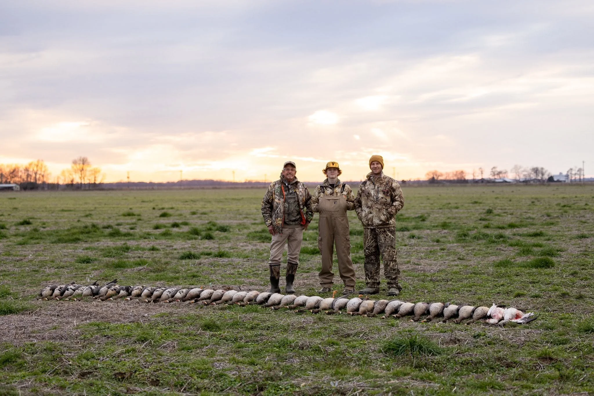 Three hunters dressed in camouflage and hunting gear standing in a field at sunrise. In front of them is a row of freshly shot geese laid out on the ground.