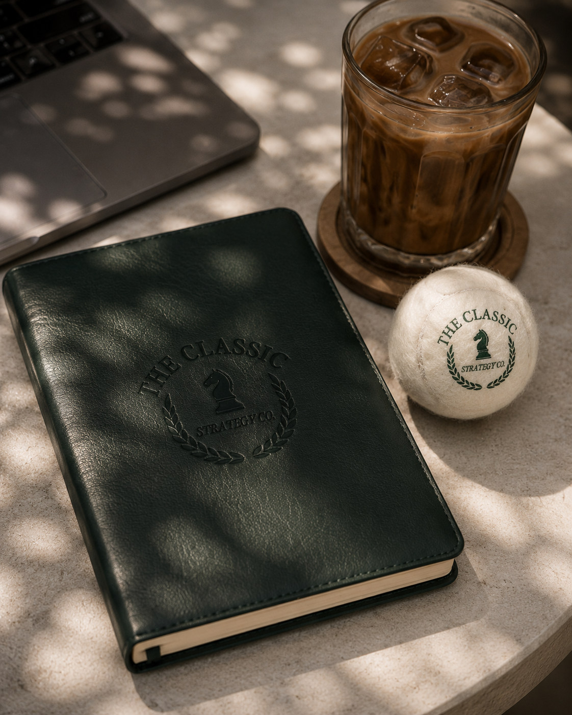 A black leather notebook with the words 'The Classic Strategy Co.' embossed on it, a tennis ball with the same logo, a glass of iced coffee on a wooden coaster, and a partially visible laptop on a light-colored table with dappled sunlight.